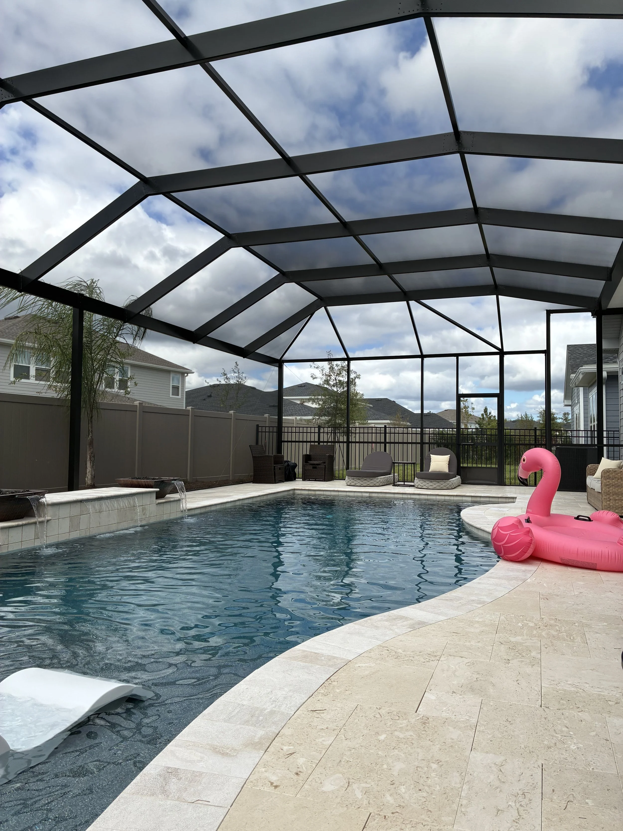 A screened outdoor patio enclosure with a clear roof, surrounded by a black metal frame. The area has a white tile floor, and there is a view of a grassy yard with trees and a fence in the background.