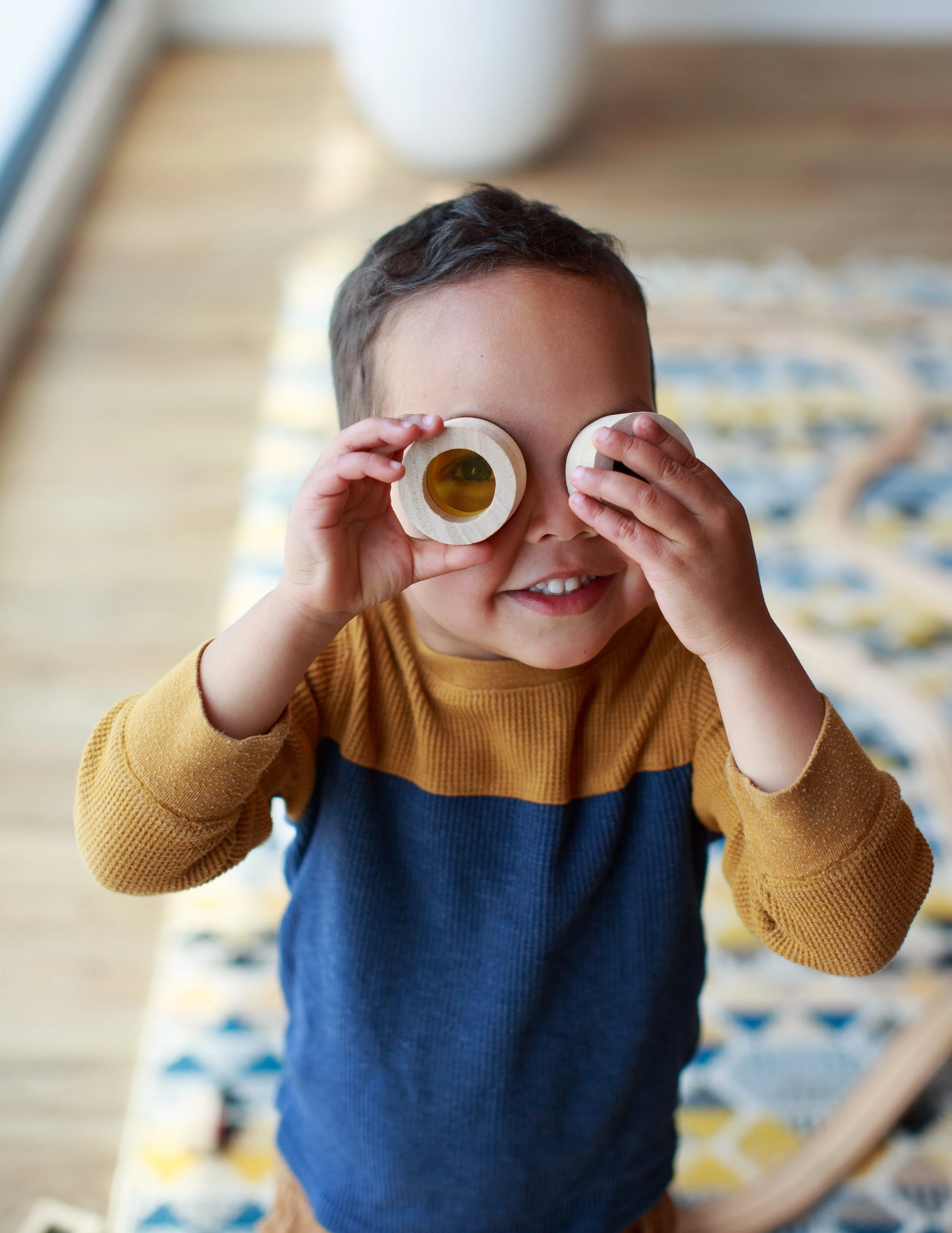 A young boy holding wooden toy binoculars up to his eyes, smiling, in a room with a patterned rug and a large window.