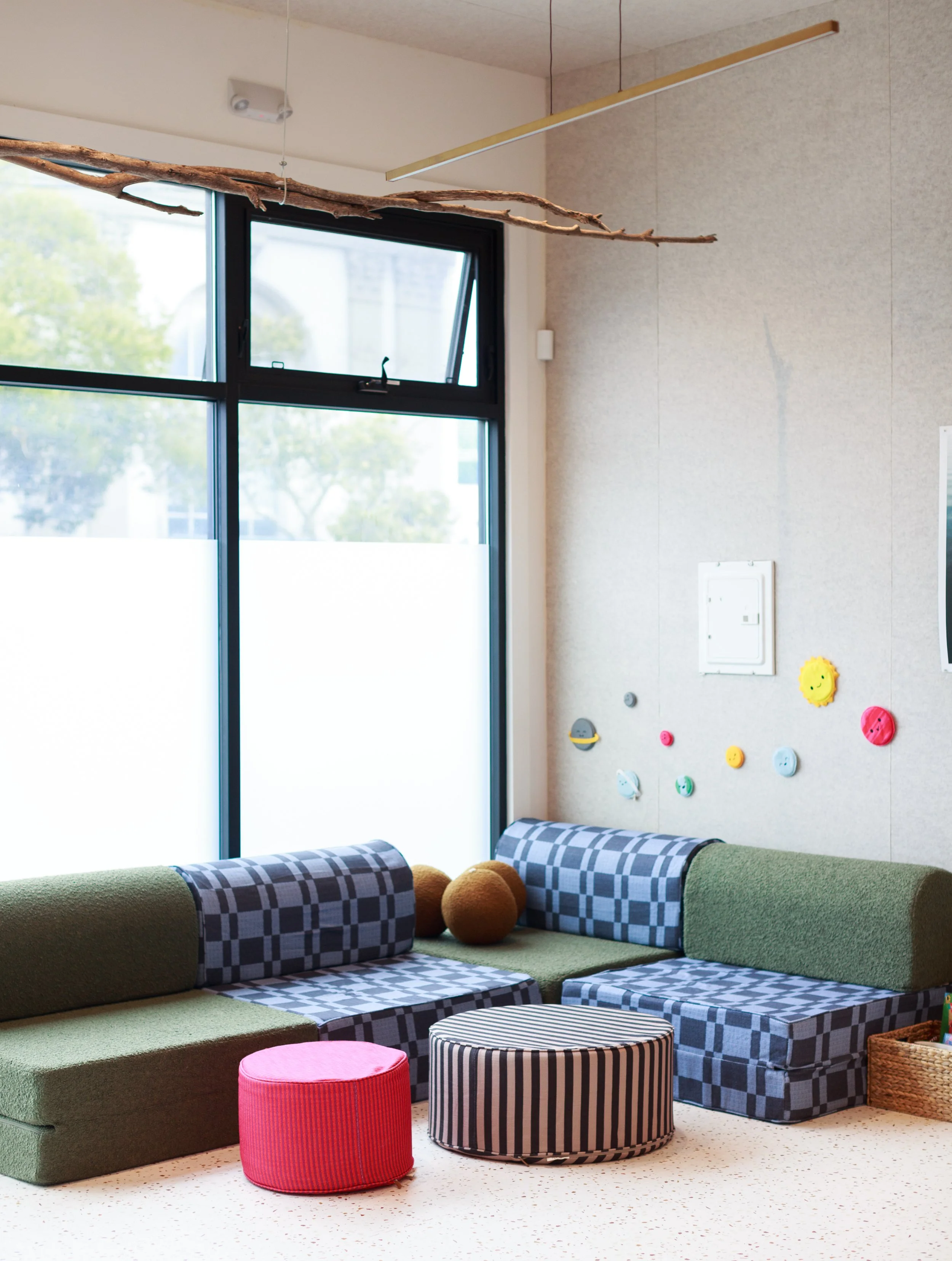 Colorful playroom with modular sofa, striped and checkered ottomans, and wall decorations, next to large window with trees outside.