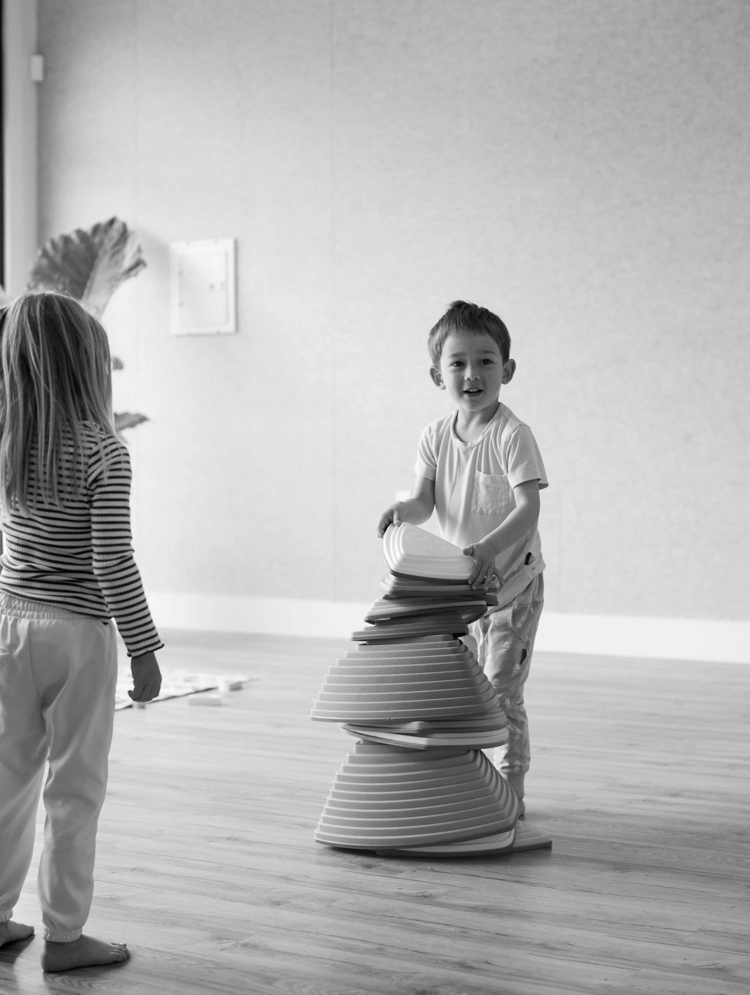 Two children, a boy and a girl, playing with large foam puzzle pieces indoors. The boy is stacking the pieces, and the girl is watching.