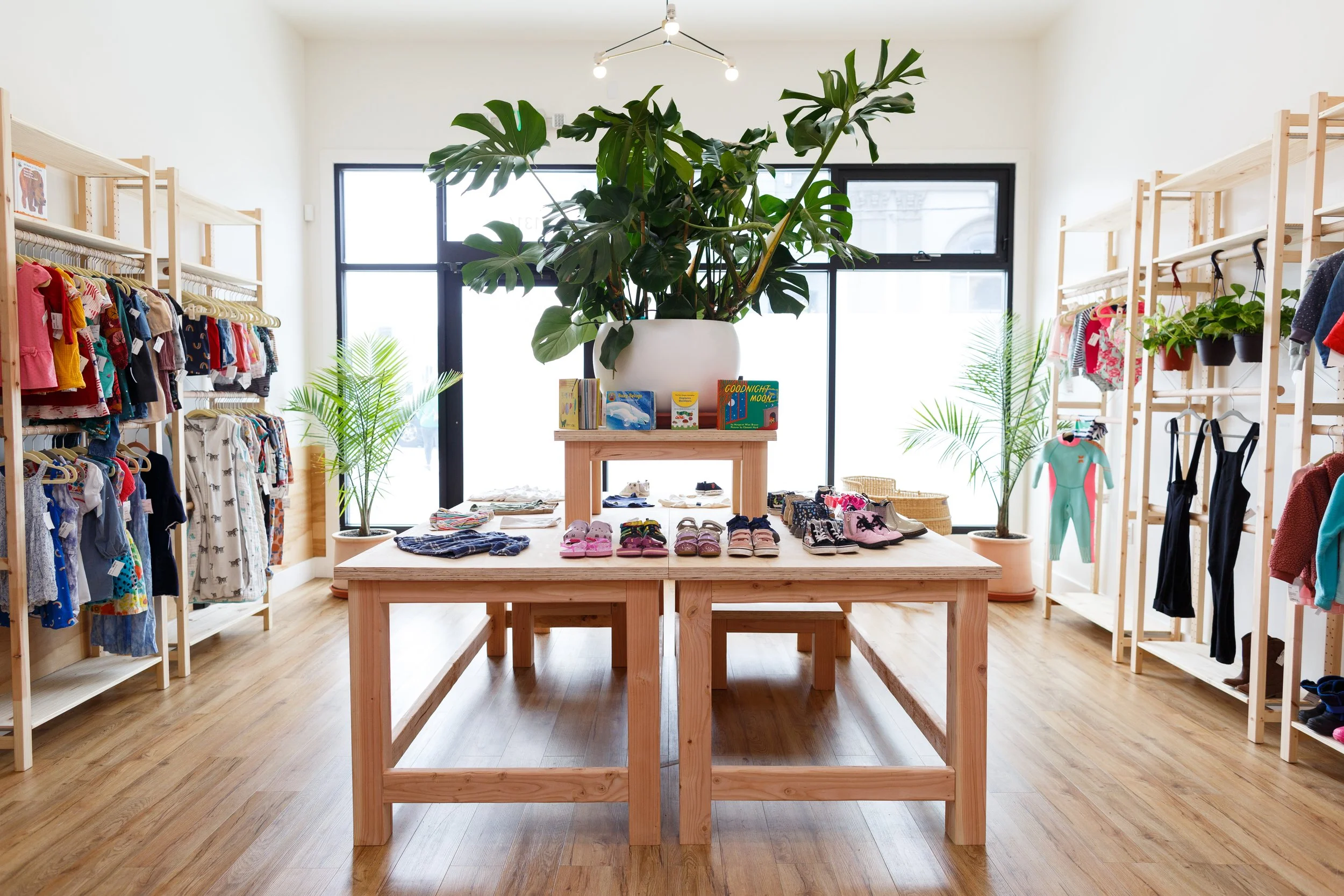 Children's clothing store with wooden shelves and tables, displaying clothes, shoes, and books. Large potted plants and a big window in the background.
