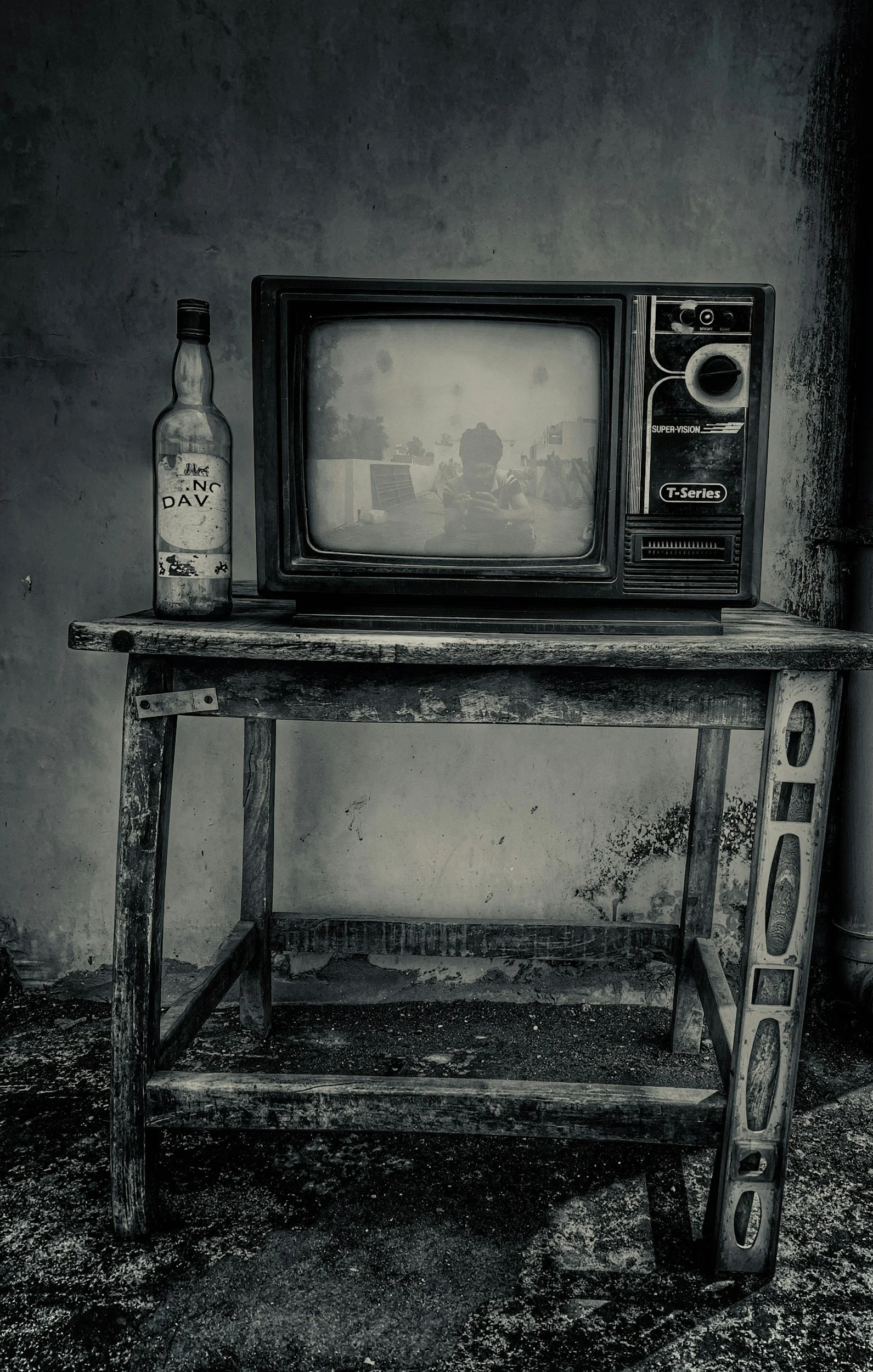 An old, black-and-white photo of a vintage television set and a bottle on a worn wooden table against a rough wall. The TV is turned off, reflecting a person taking the photo.