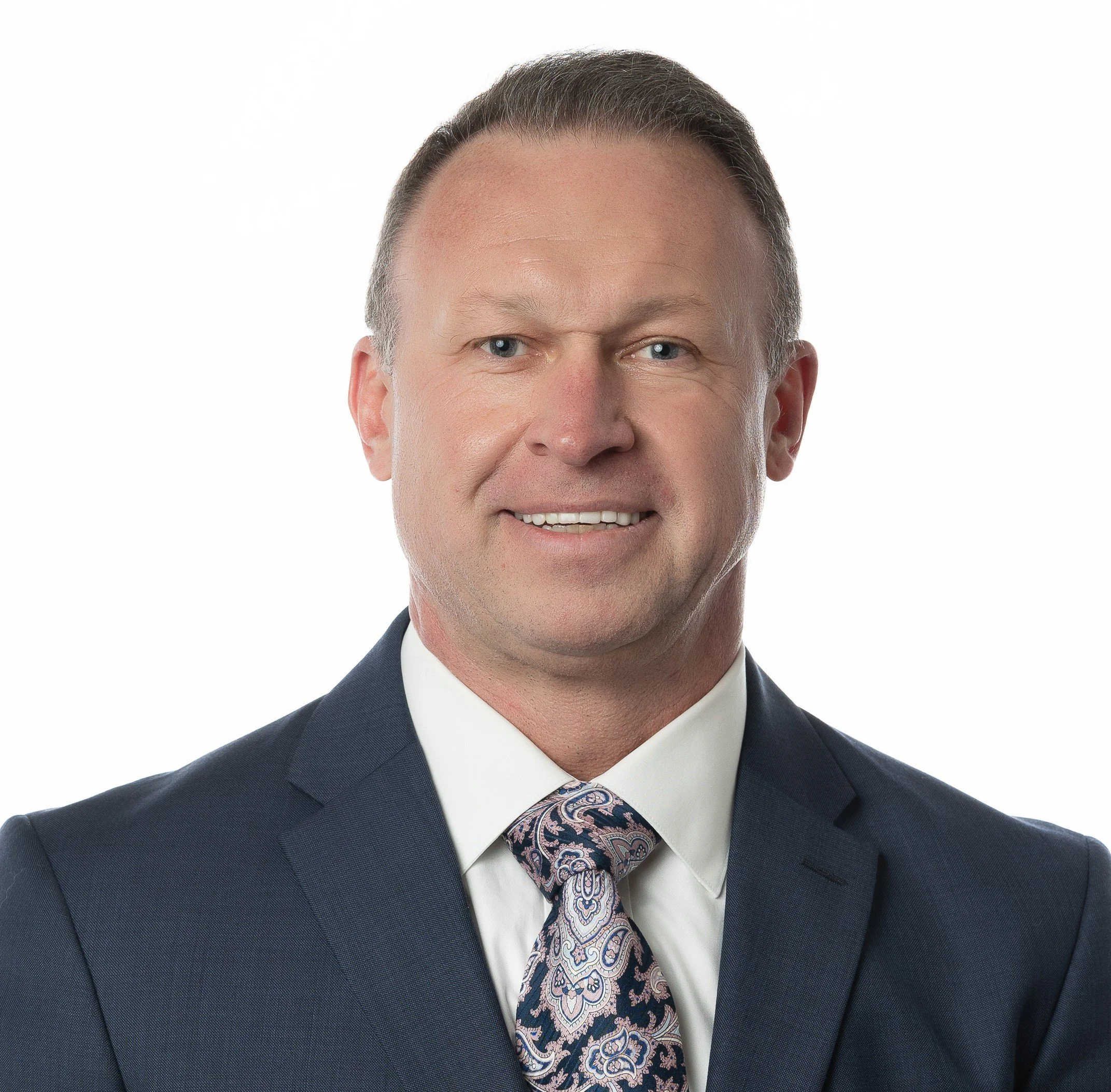 Head and shoulders portrait of a man in a dark suit, white shirt, and paisley tie, smiling against a plain white background.