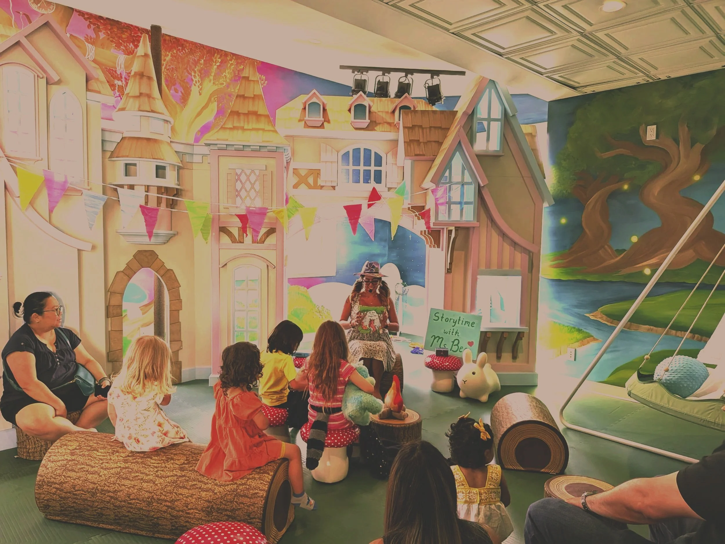 Children and adults gathered in a colorful indoor space watching a storytime session with a woman dressed as a fairy tale character. The setting features a whimsical, castle-like backdrop with murals of a tree and landscape, decorated with bunting and plush toys.