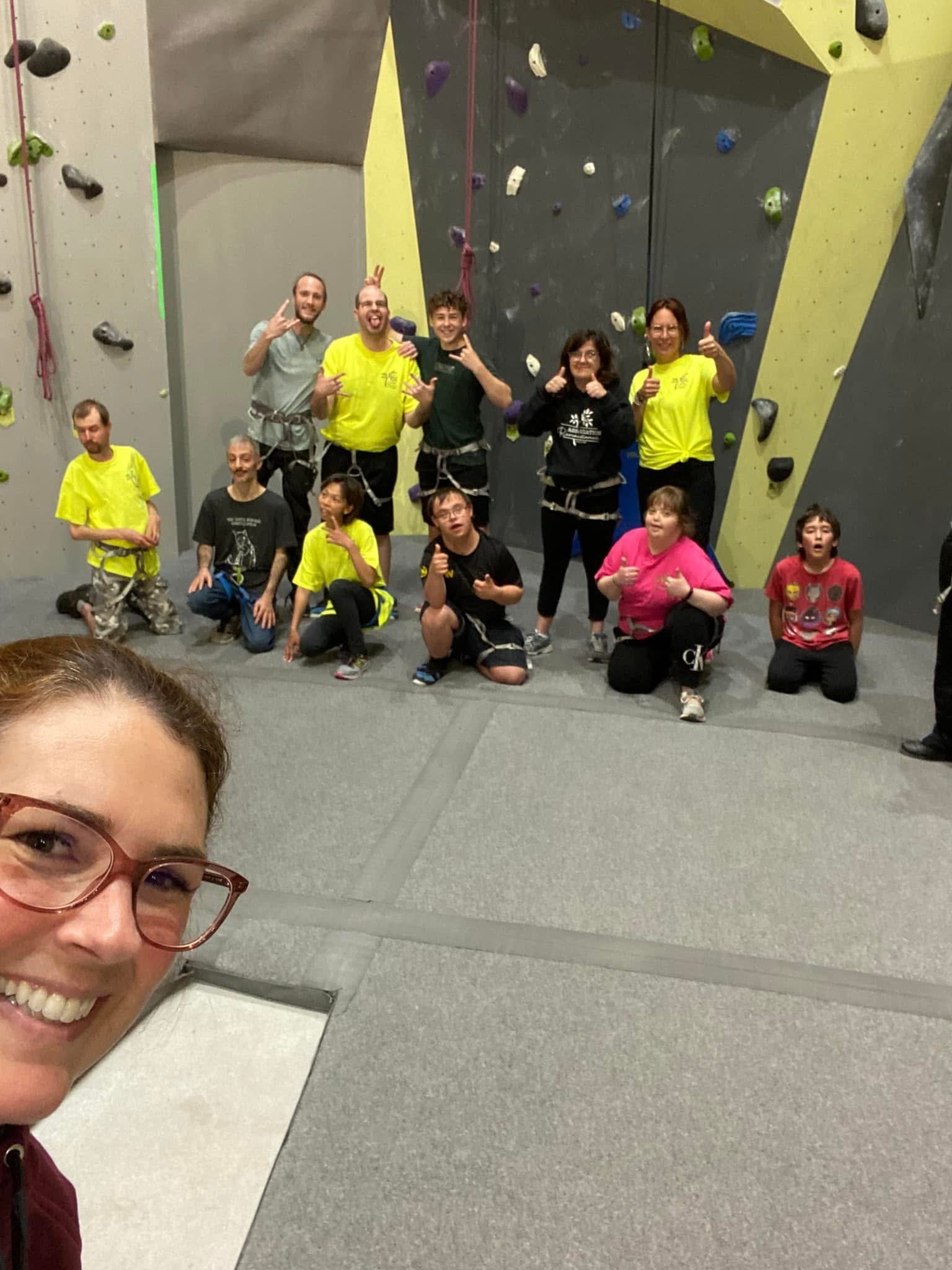Des personnes posent devant un mur d'escalade, portant des équipements de sécurité et des vêtements décontractés. Une femme est au premier plan, souriant pour un selfie.