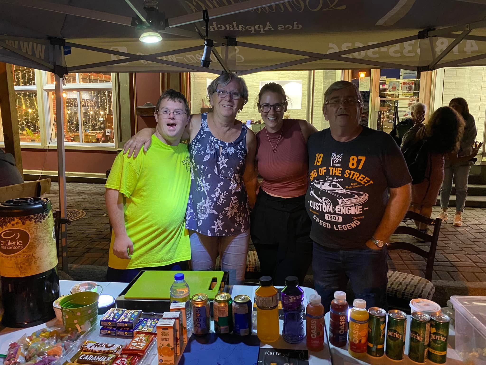 Quatre personnes souriantes debout derrière une table avec des boissons et snacks, sous une tente en soirée.
