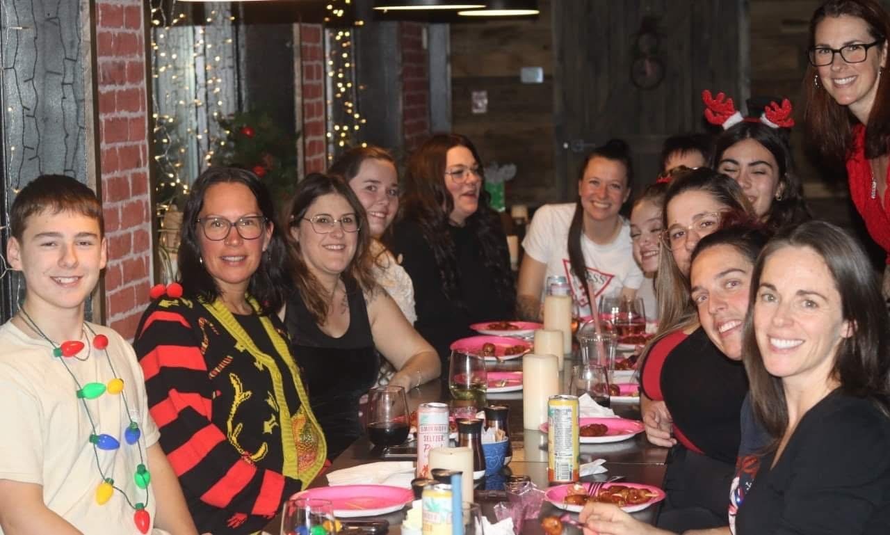 Groupe de personnes assises autour d'une table, souriantes et portant des vêtements festifs. Décorations de Noël en arrière-plan.
