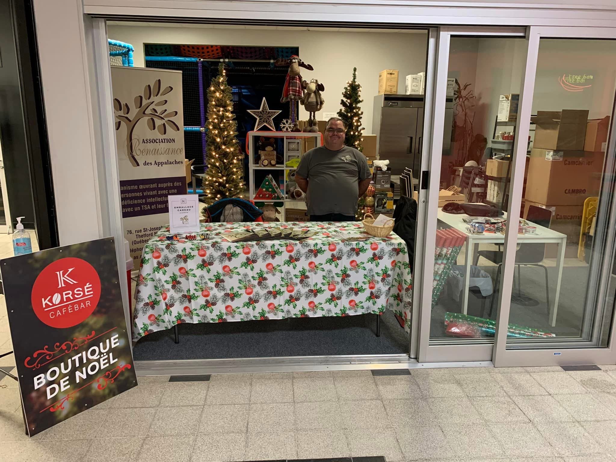 Boutique de Noël avec un stand décoré, un homme debout derrière une table avec une nappe à motifs de Noël, des affiches indiquant "Boutique de Noël" et "Association Renaissance des Appalaches", des décorations de Noël et un sapin illuminé à l'intérie