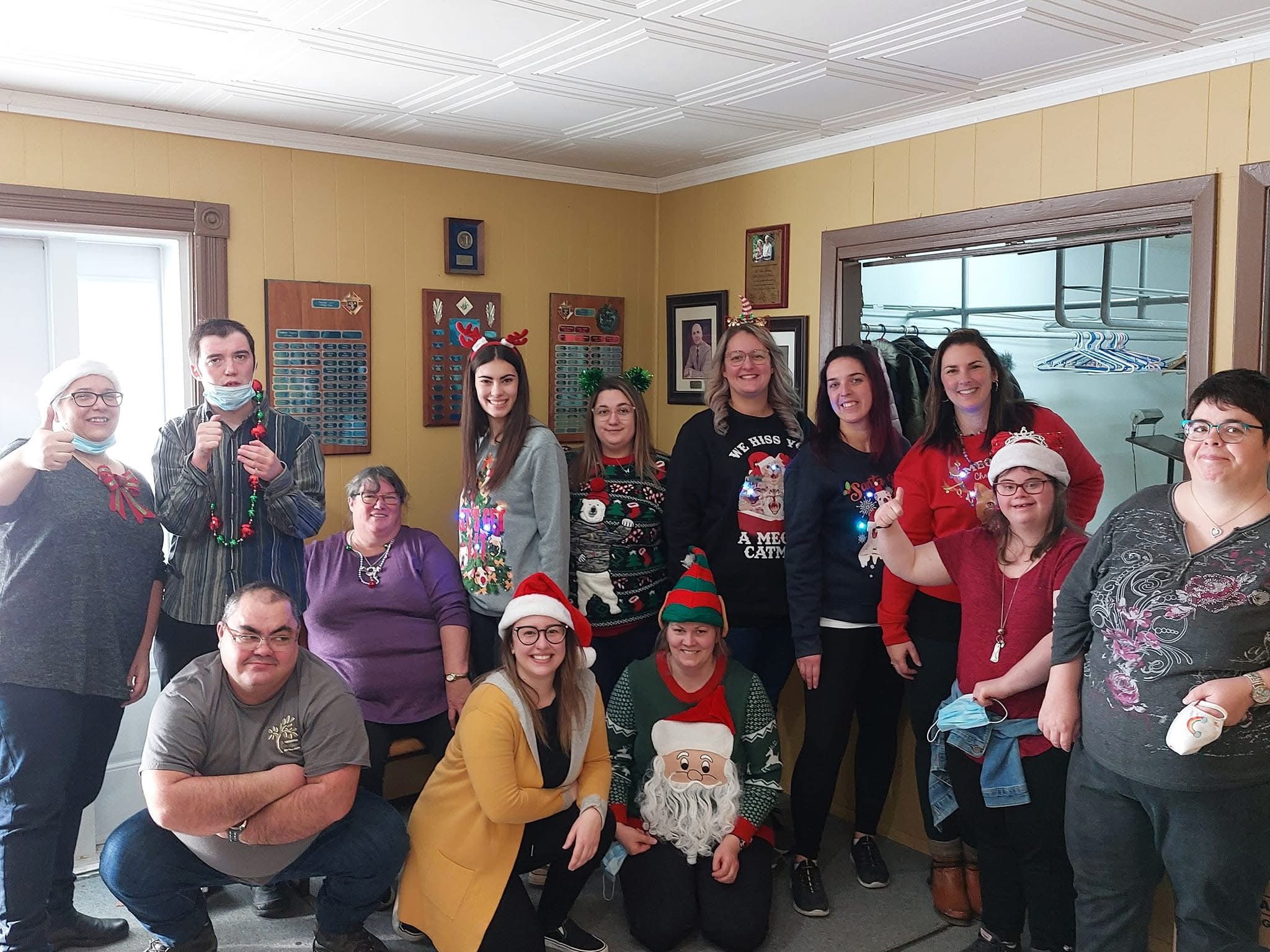 Groupe de personnes posant dans une pièce décorée de Noël, certaines portant des pull-overs et accessoires festifs. Il y a divers éléments de décoration sur les murs et certains membres du groupe tiennent des objets en lien avec les fêtes.