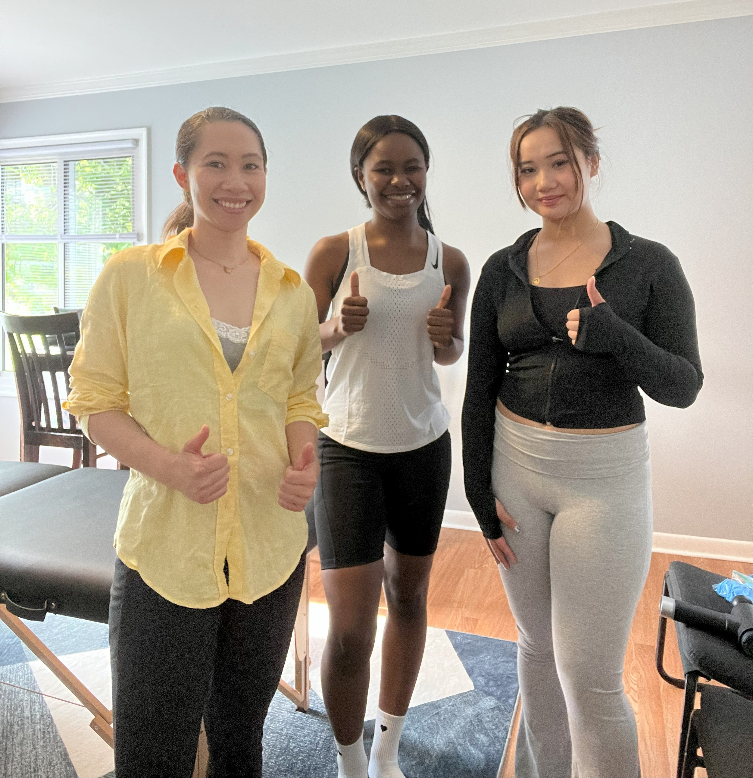 Three women standing together indoors, smiling, and giving thumbs-up gestures, with a massage table and chairs in the background.