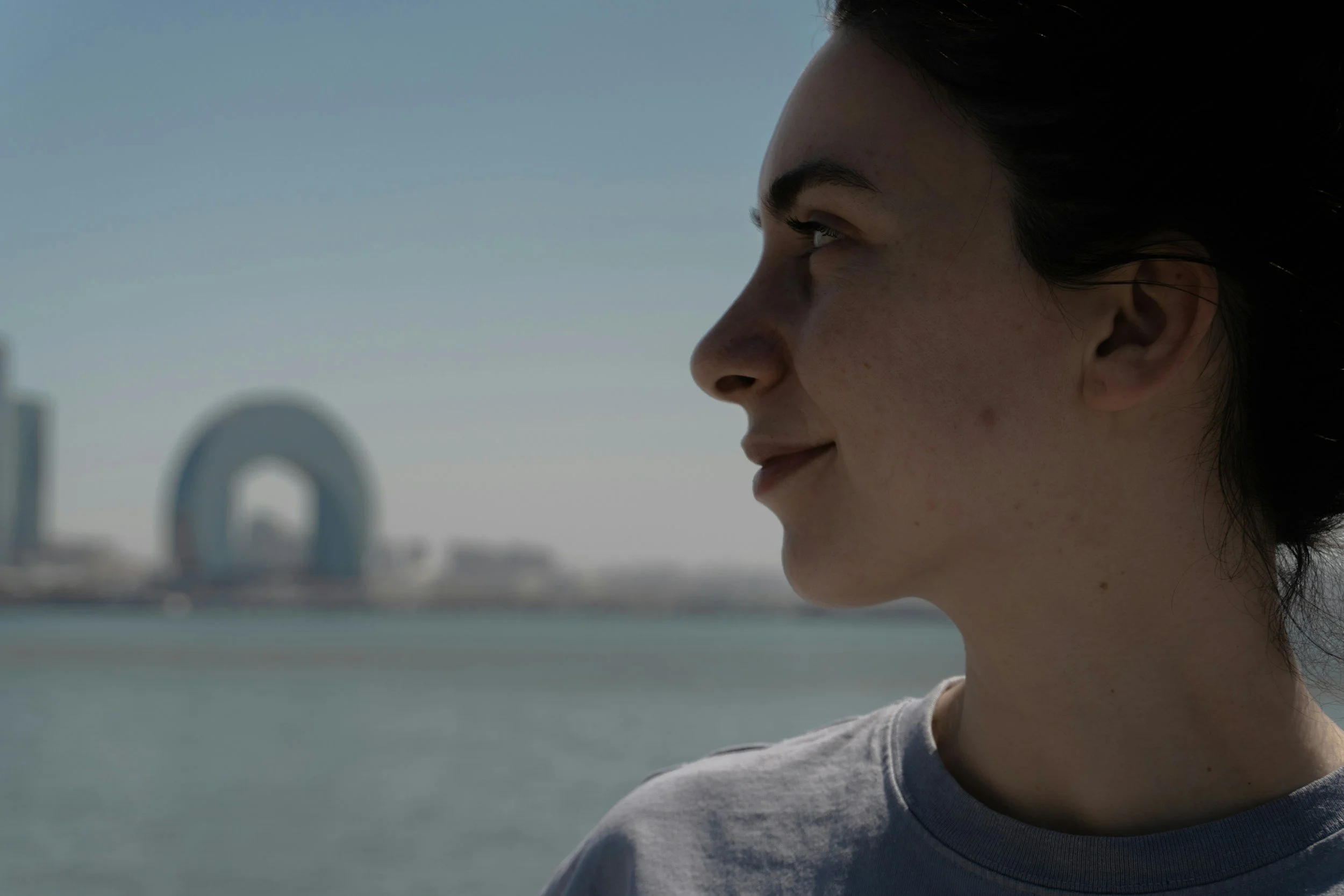 Side profile of a smiling woman with dark hair and freckled skin, outdoors near a body of water with a blurred modern city skyline, including a distinctive circular building, in the background.