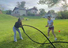 Two women playing tug of war with a thick black rope on a grassy yard, with a house and trees in the background.