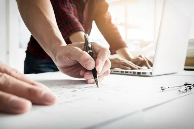 Two people working at a table with one person signing a document and the other person typing on a laptop.