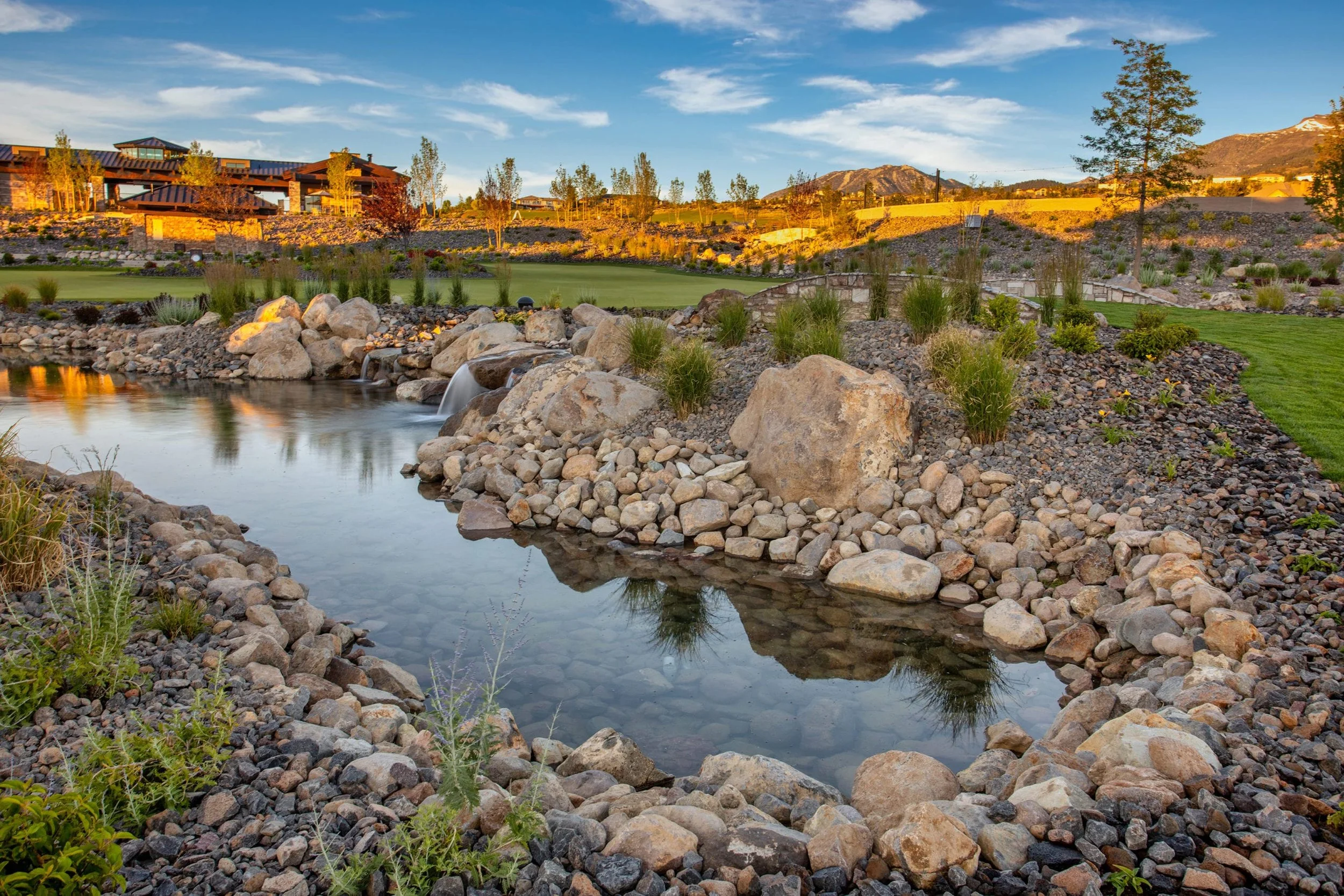 ArrowCreek A landscaped water feature with rocks and small plants, a small waterfall, in a residential area with modern houses and mountains in the background, under a partly cloudy sky.