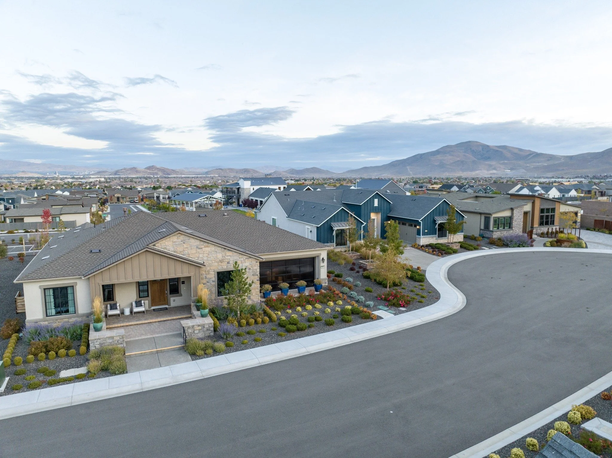 Caramella Ranch Aerial view of a suburban neighborhood with modern houses, landscaped yards, and a curved street, with mountains in the background under a cloudy sky.