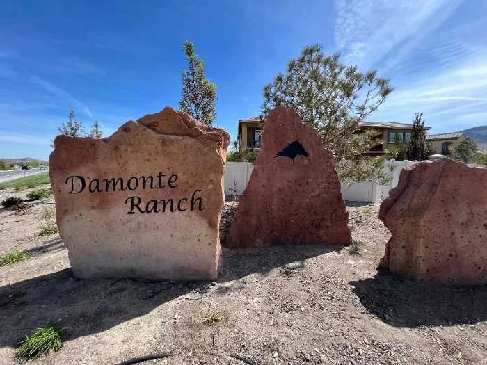 Three large rocks at the entrance of Damonte Ranch, with one displaying the text 'Damonte Ranch' and another with a silhouette of a bird, possibly a raven, on it. There are trees and buildings in the background under a bright blue sky.