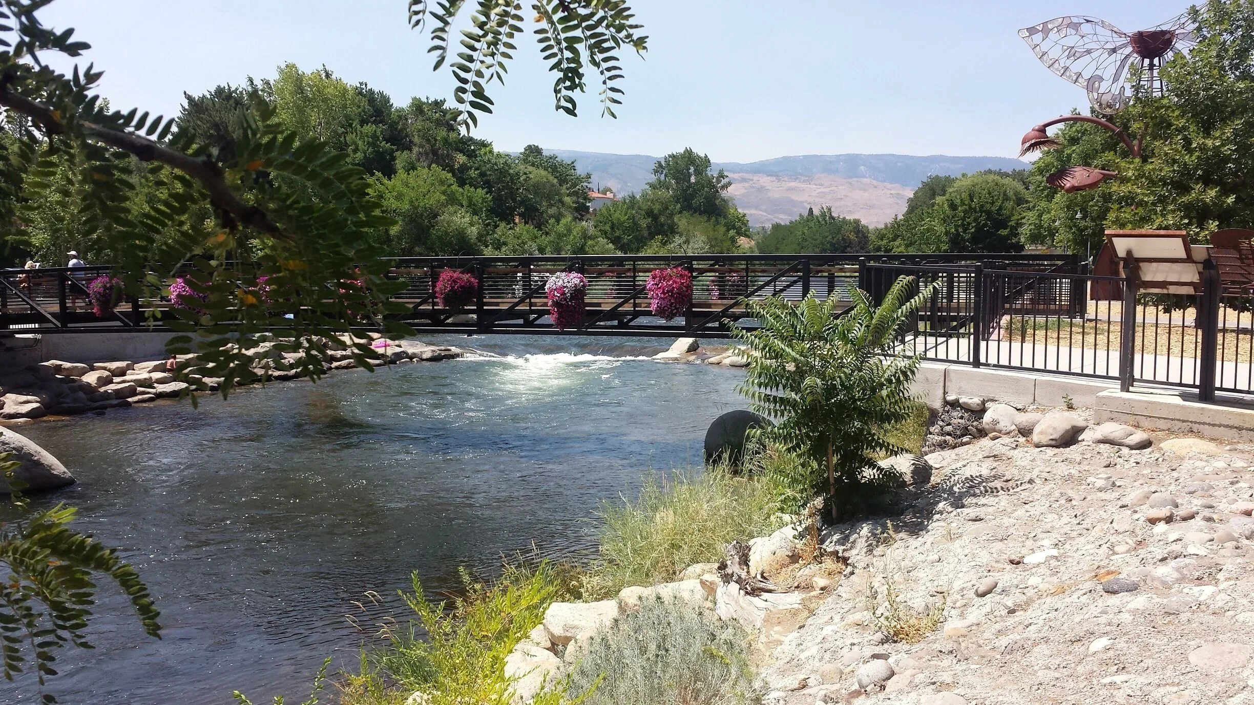 Old Southwest Reno A small river with a black metal bridge decorated with pink and purple flowers, surrounded by green trees and mountains in the background.