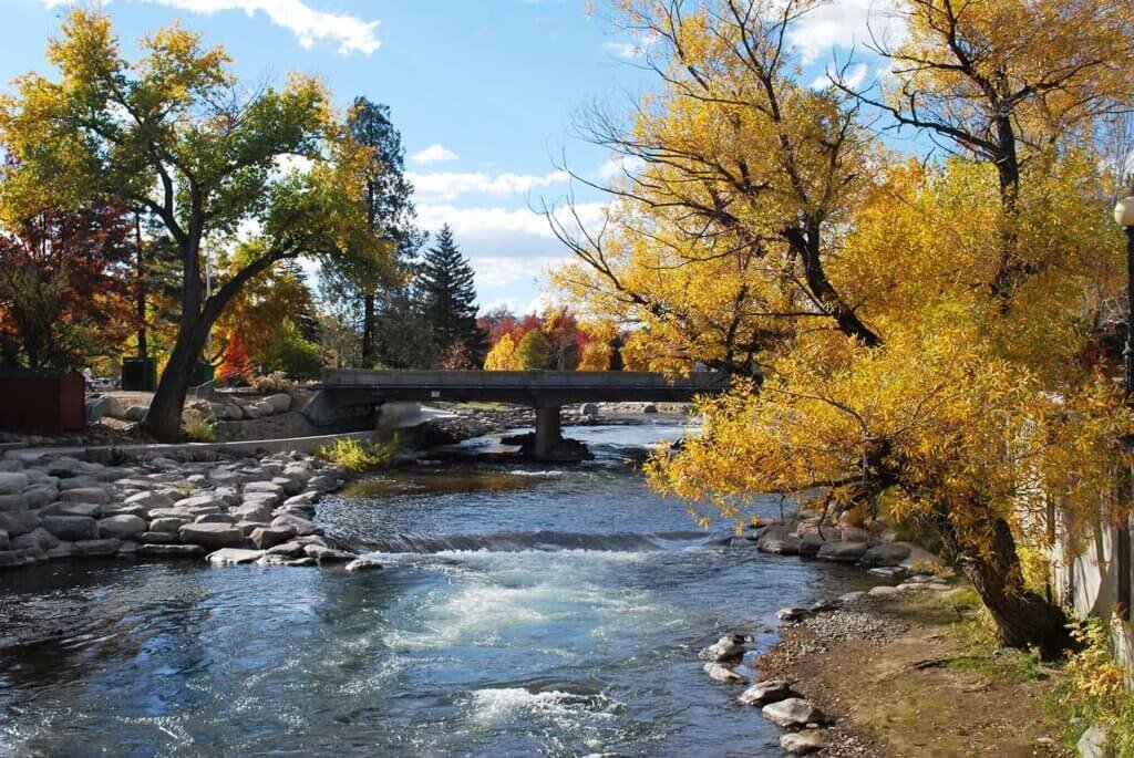 Verdi A river running through a park with trees showing fall foliage, a bridge crossing the river, and rocks along the riverbank under a partly cloudy sky.
