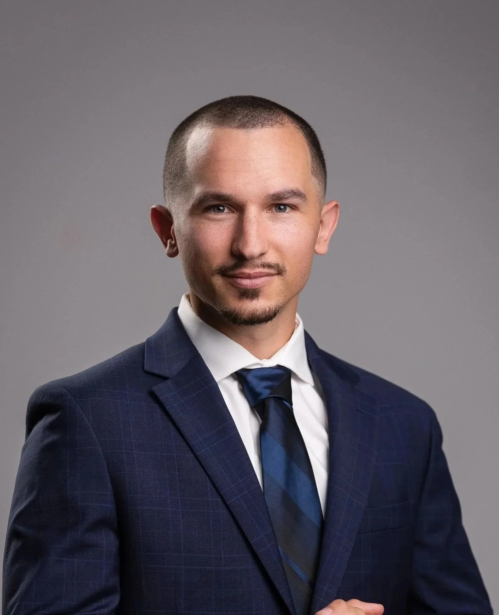 A man in a navy blue suit with a white shirt and matching navy blue tie, posing against a gray background.