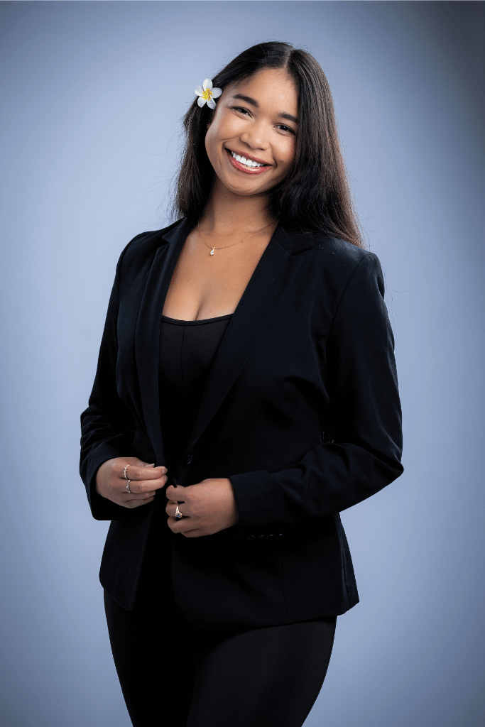 A young woman from Landeck Real Estate Team with long dark hair and a white flower behind her ear, smiling, wearing a black blazer and black top, standing against a light blue background.