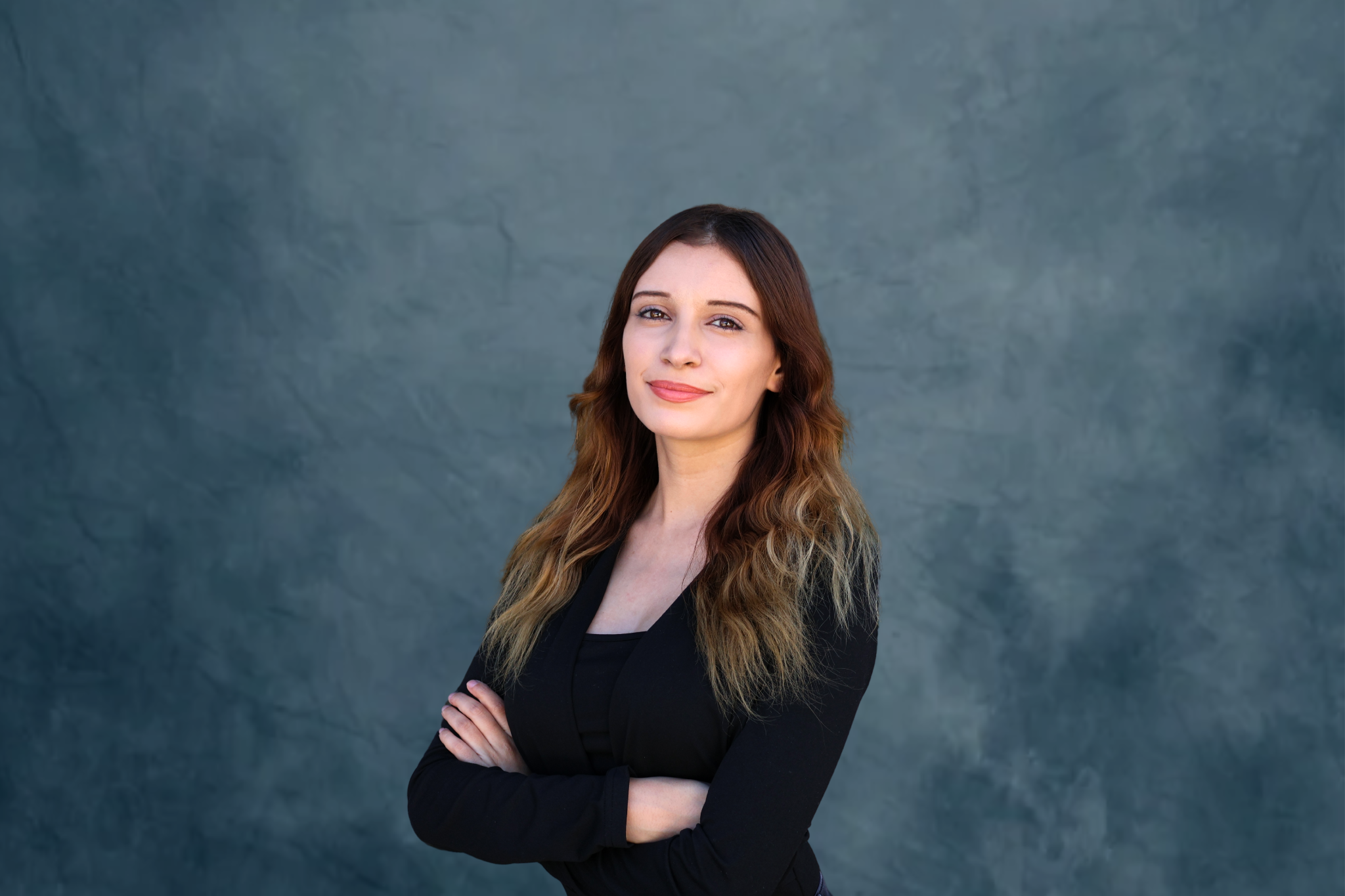 A confident woman with light skin and long, wavy brown hair standing with her arms crossed against a textured blue-gray background.