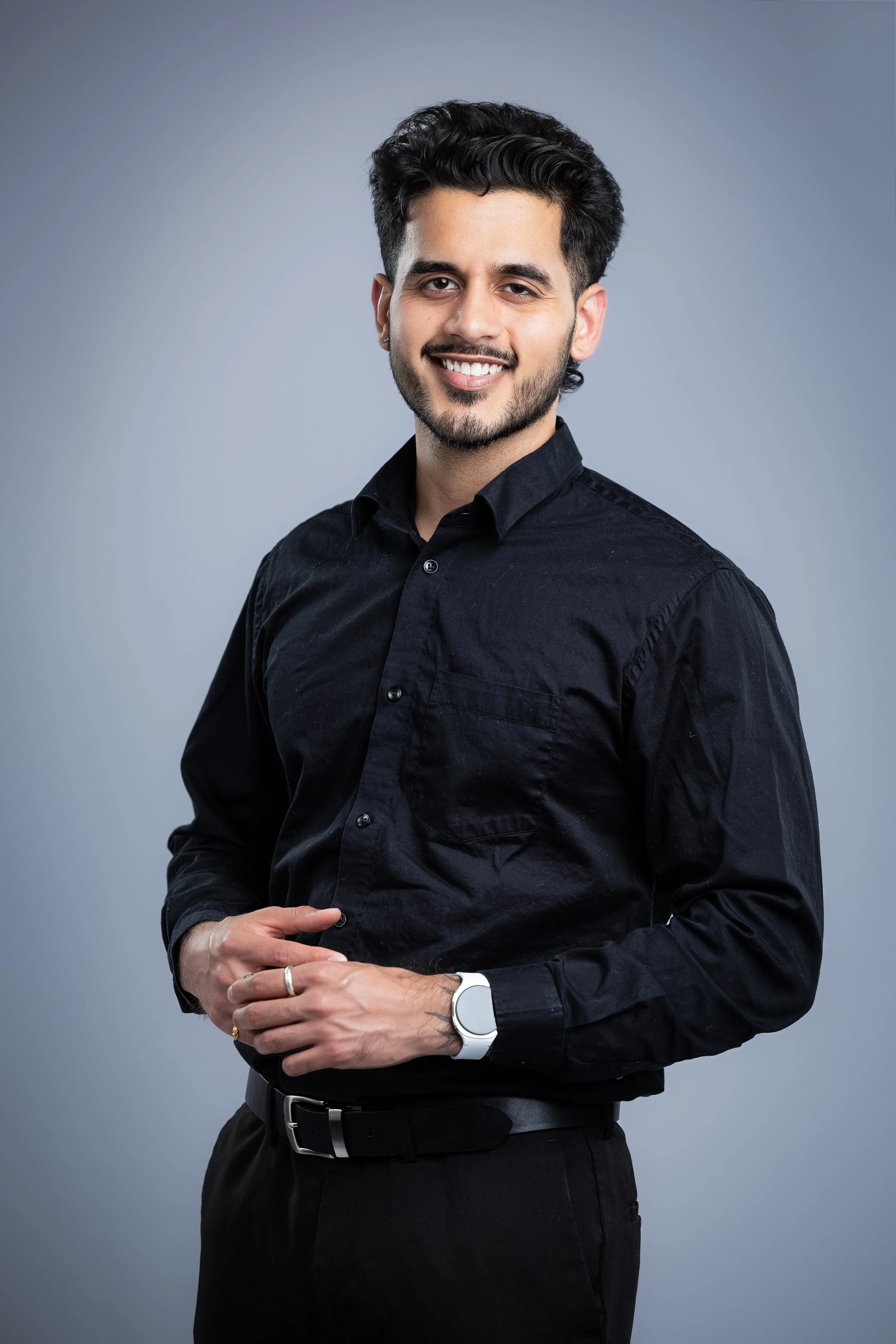A young man with dark hair and a beard, smiling, wearing a black dress shirt, standing against a gray background.