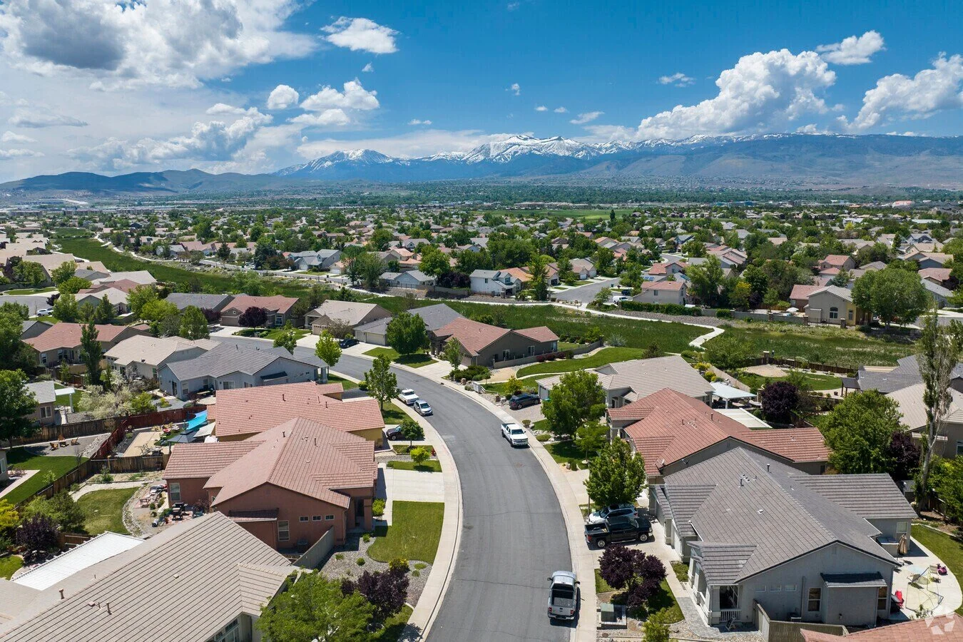 Double Diamond Aerial view of a suburban neighborhood with houses, trees, and cars on the street, mountains with snow in the background, under a partly cloudy sky.