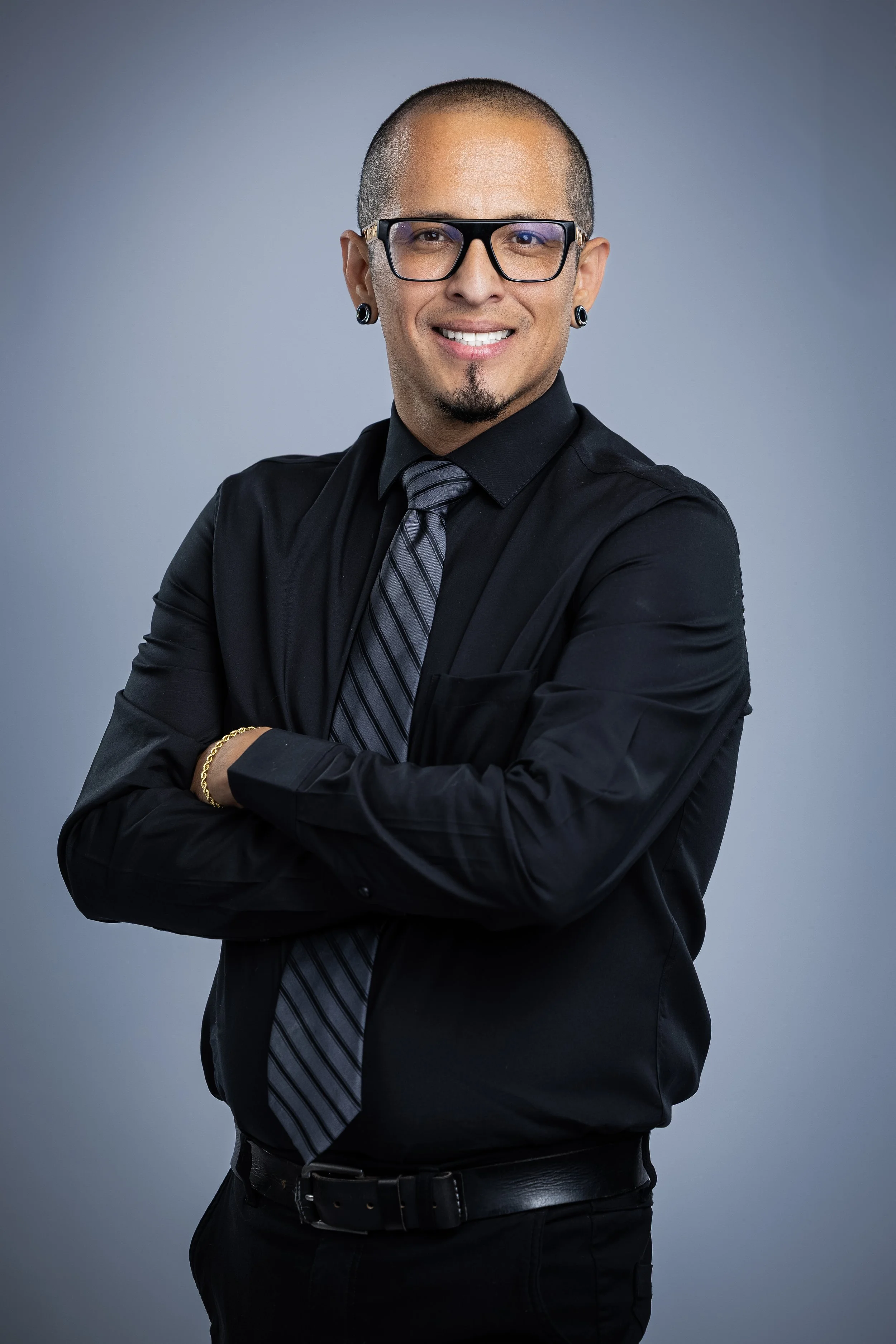 Man with glasses and earrings wearing a black dress shirt and a striped tie, standing against a gray background with arms crossed, smiling