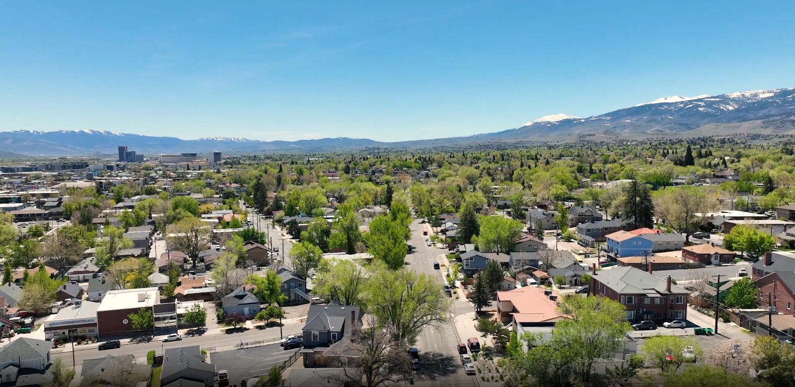 Midtown Reno Aerial view of a suburban neighborhood with green trees, houses, and streets, with mountains in the distance under a clear blue sky.