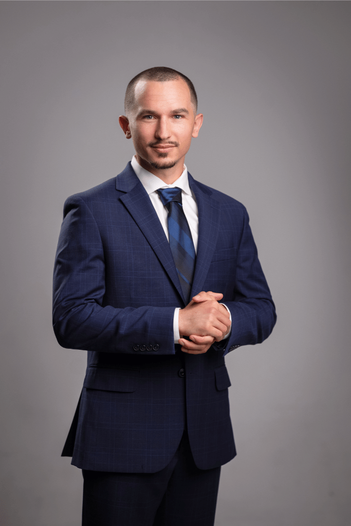 Alex Landeck, Landeck Real Estate Team, in a dark blue checkered suit with a white shirt and blue tie, standing with his hands clasped in front of him against a plain gray background.