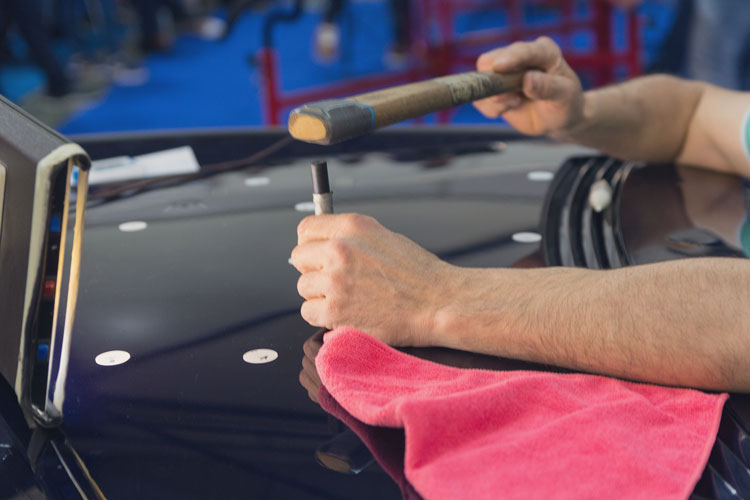 Person using a hammer and tool on a car's hood with a red cloth nearby.