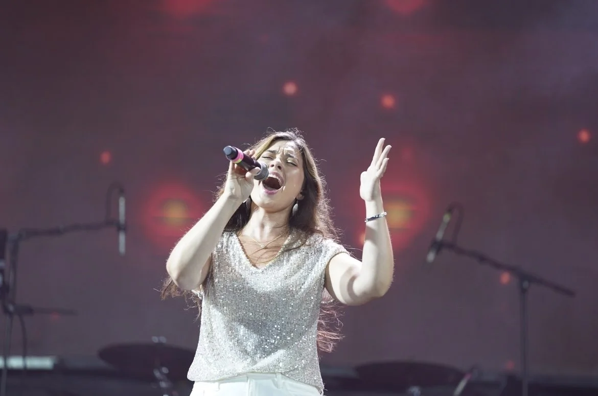 A woman singing passionately into a microphone on stage with a dark background and red stage lights.