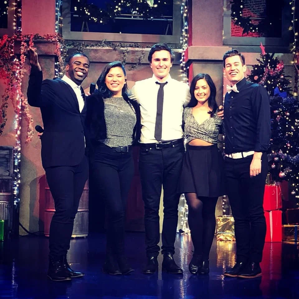 Group of five young adults dressed in formal attire, standing together at a festive Christmas party, with a decorated Christmas tree and wrapped presents in the background.
