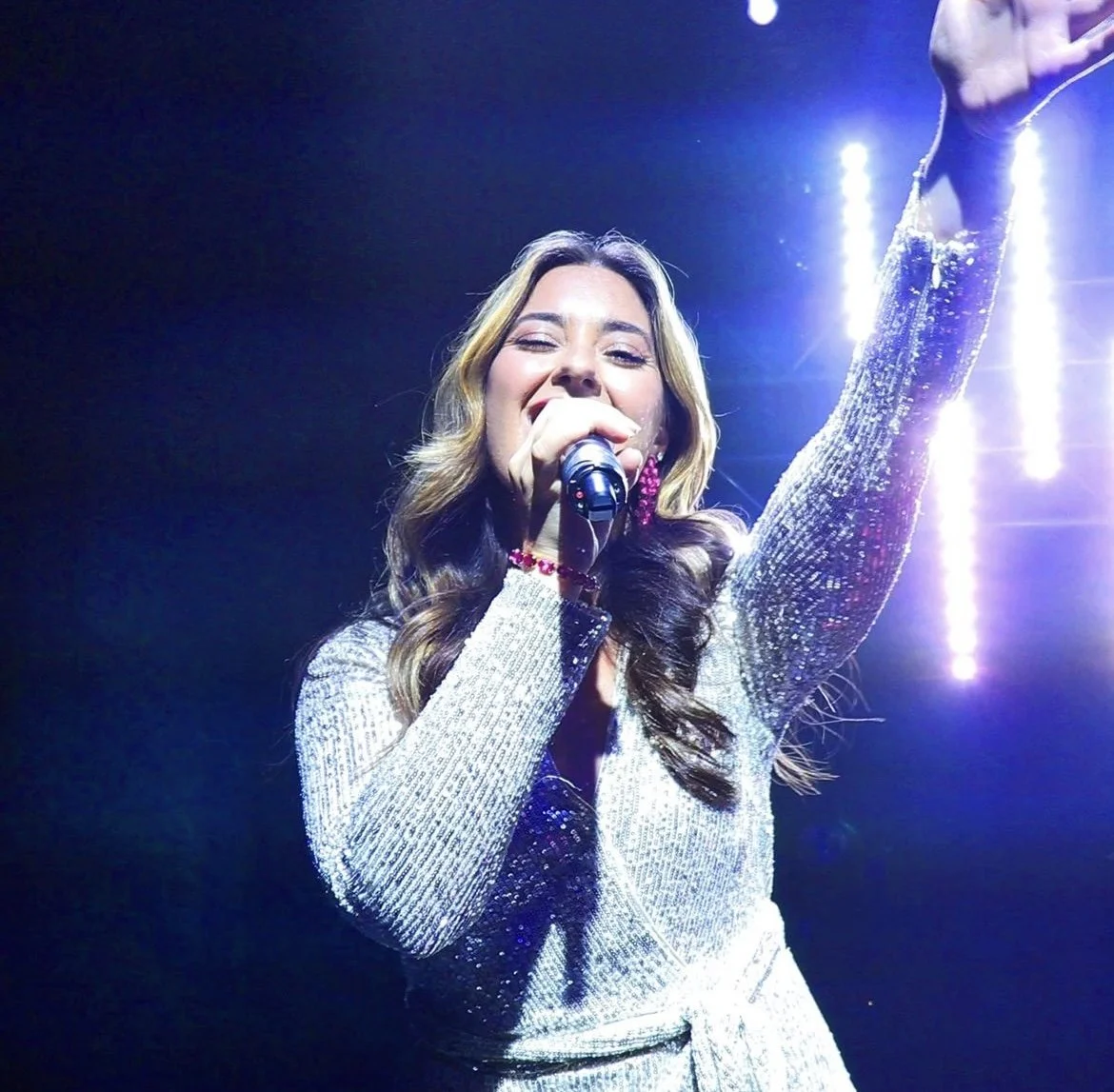 A woman singing into a microphone on stage, wearing a shiny silver dress with long sleeves and a pink beaded bracelet, with bright stage lights in the background.