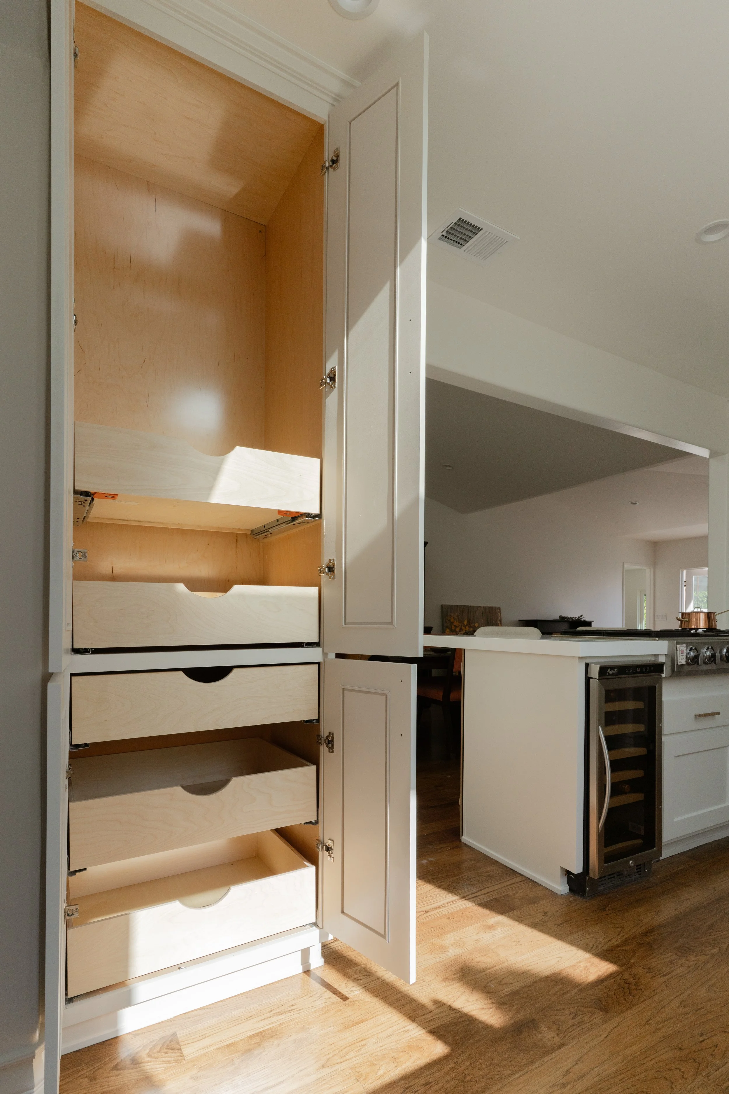 Open kitchen cabinet with multiple empty drawers and a built-in wine cooler. Part of kitchen counter and living area visible.