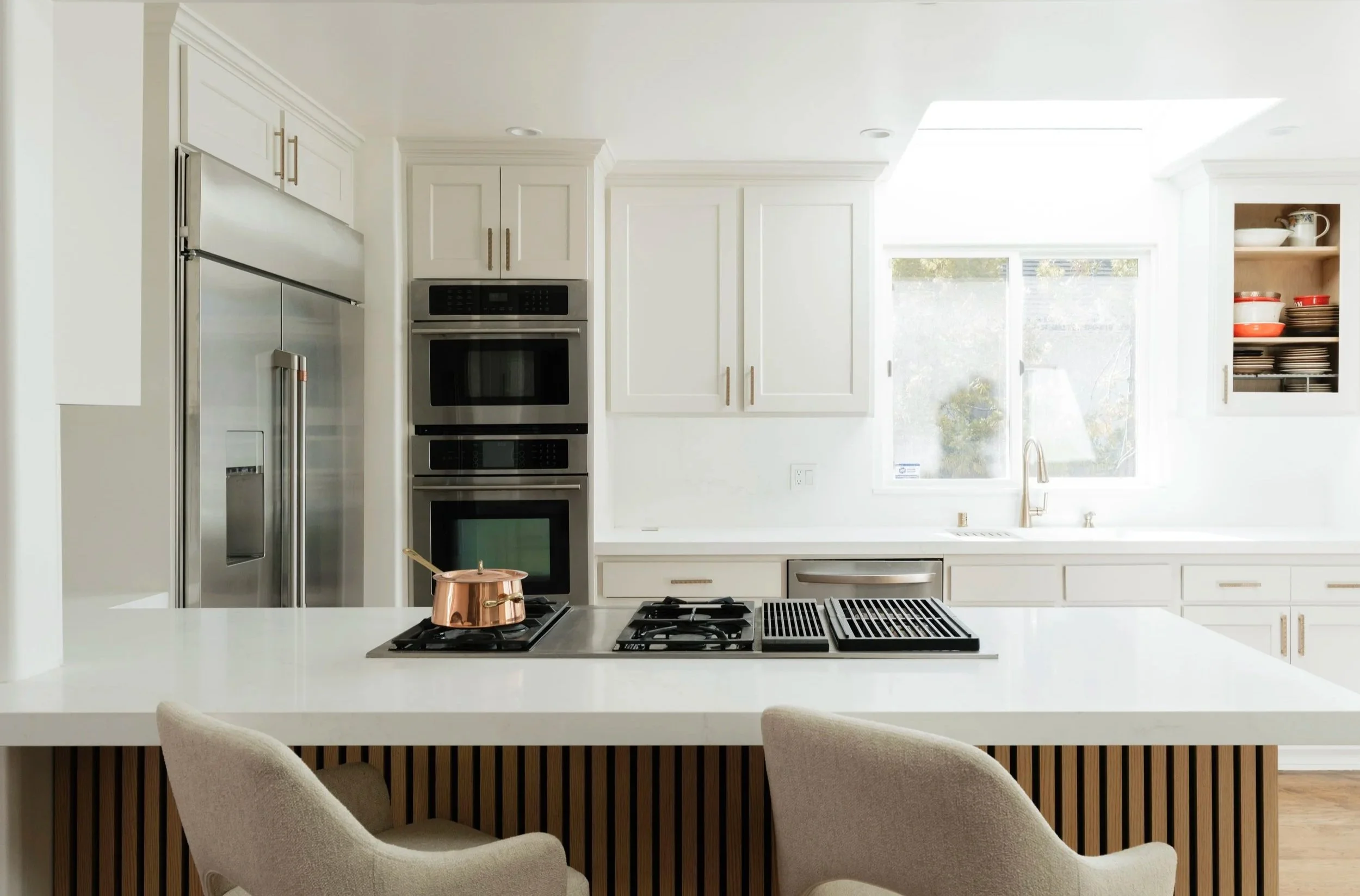 Modern kitchen with white cabinetry, stainless steel appliances, a large window, and a kitchen island with beige chairs.