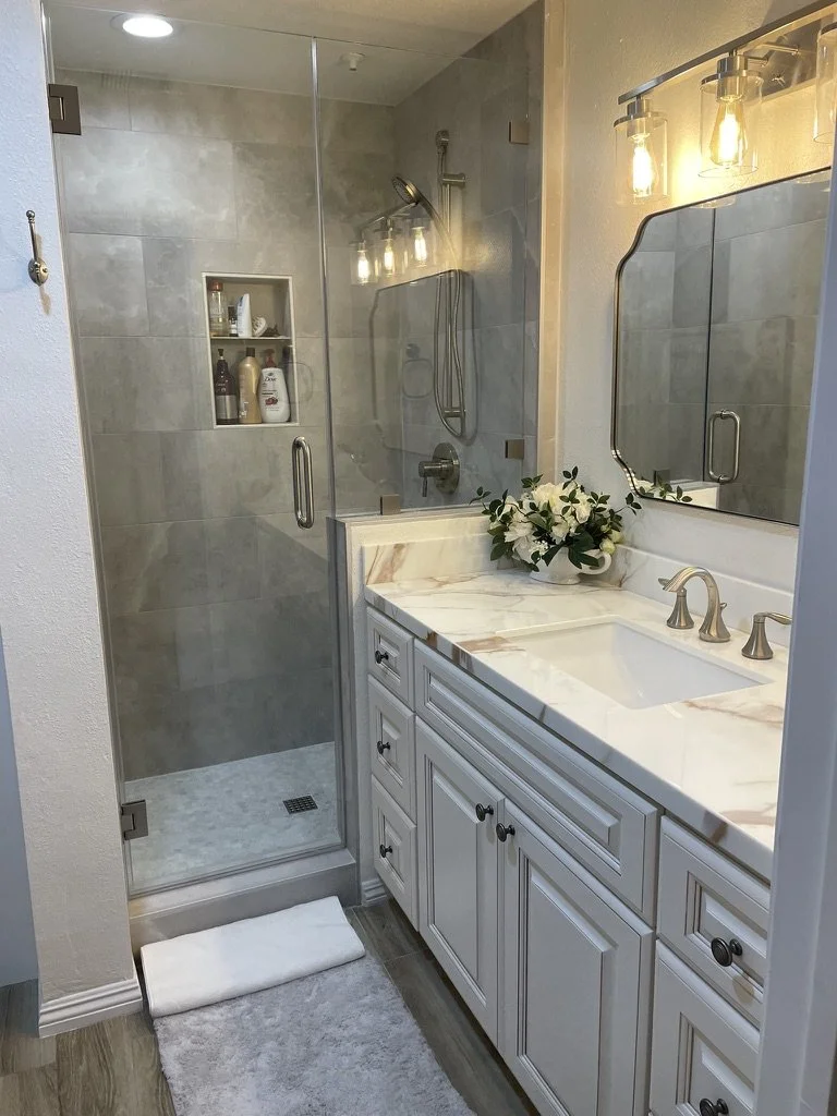 A small bathroom with a glass shower enclosure, a white vanity with a marble countertop, and a rectangular mirror. A flower arrangement decorates the countertop. The shower has built-in shelves with toiletries, and there is a rug in front of the show