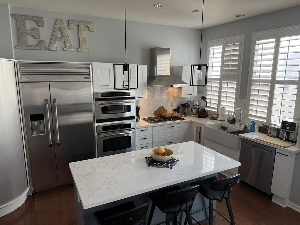 Modern white kitchen with stainless steel appliances, a white island with a fruit bowl, and large windows with plantation shutters.