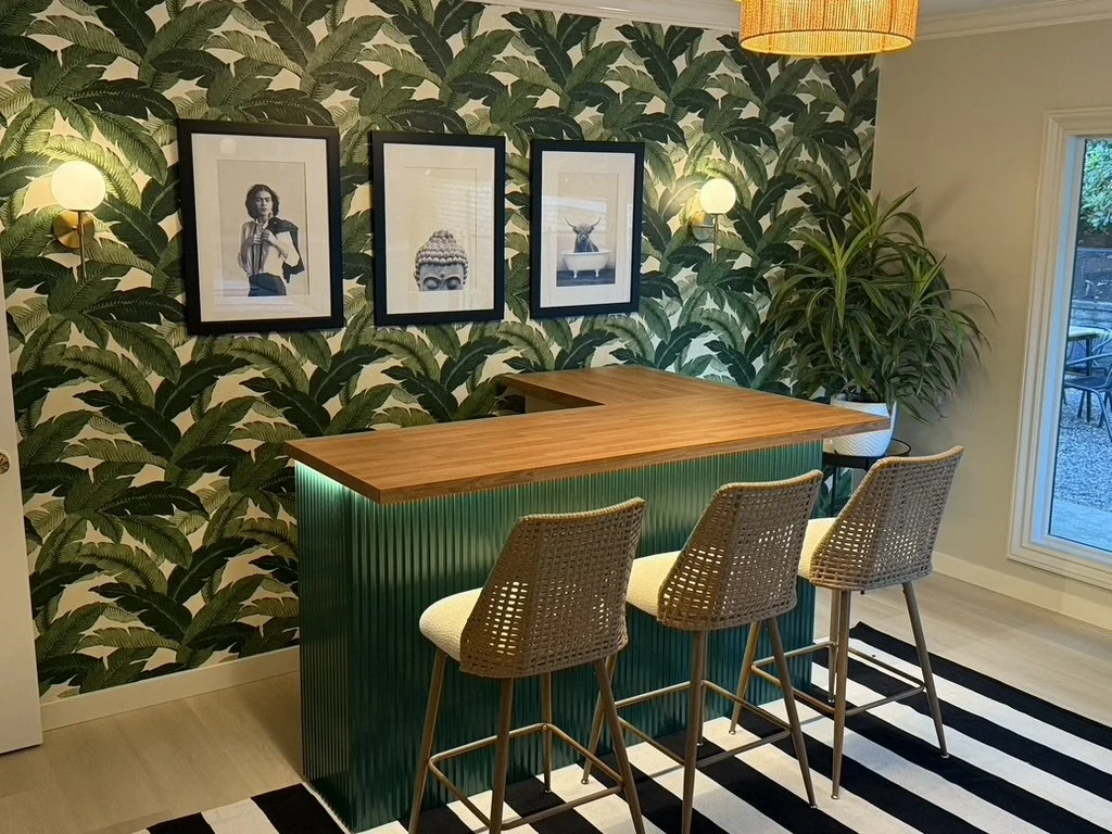 A bar area with a wooden countertop, three woven bar stools, and a green ribbed base. Behind the bar, there is a wall with a leafy green wallpaper and three framed black-and-white photos. A large potted plant is in the corner, and there is a sliding 