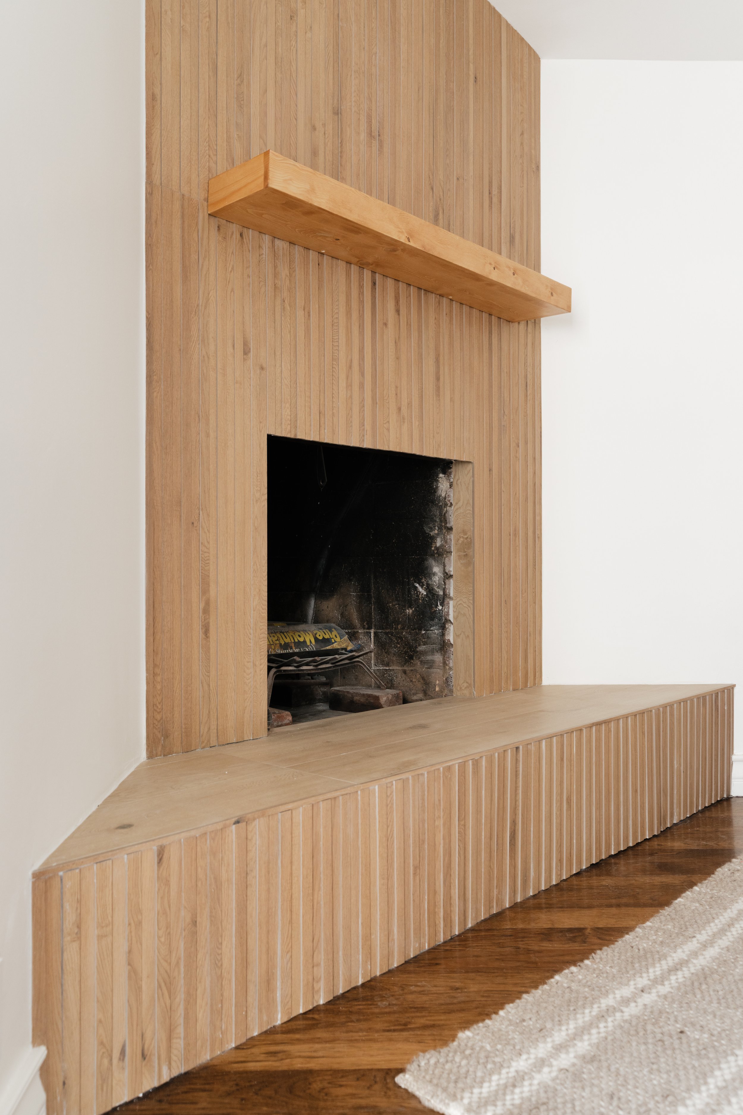 Interior view of a fireplace with a wooden mantel and a built-in wood storage bench, with a white wall on the left and a wooden floor.