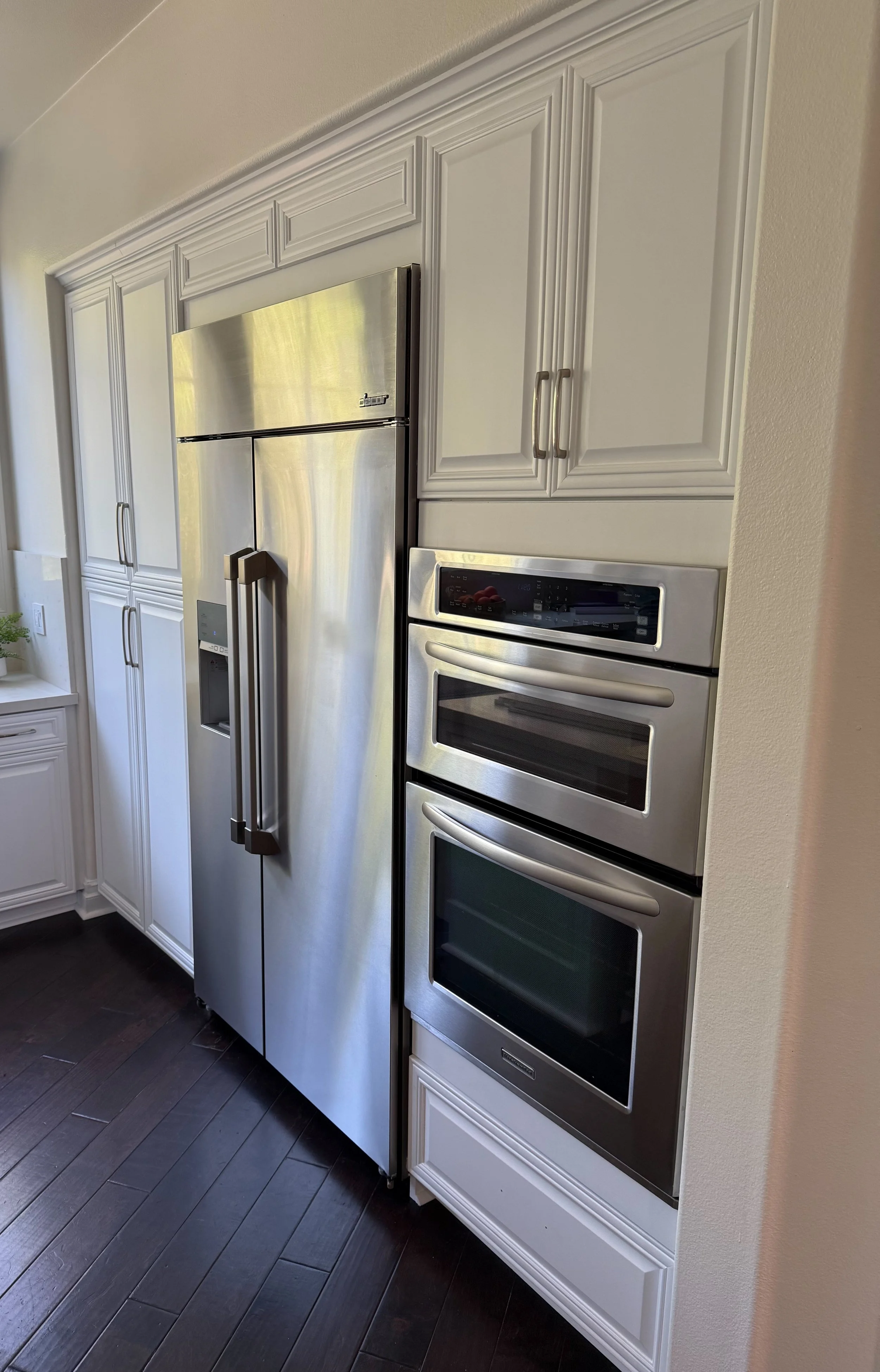 Stainless steel refrigerator and double wall oven built into white kitchen cabinets with dark hardwood flooring.