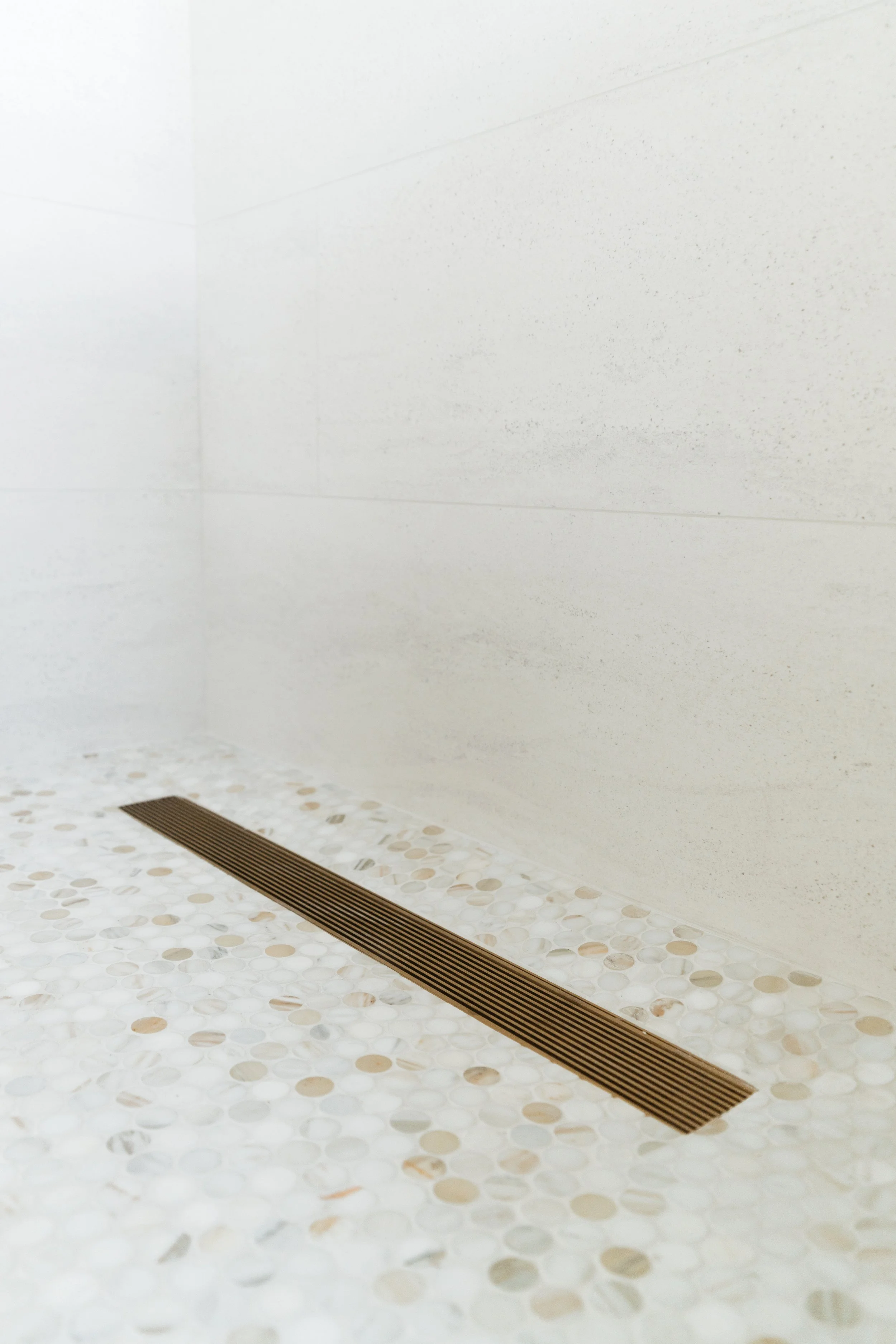 Close-up of bathroom floor tile with a champagne bronze linear drain and mosaic tile pattern, near plain white tiled walls.