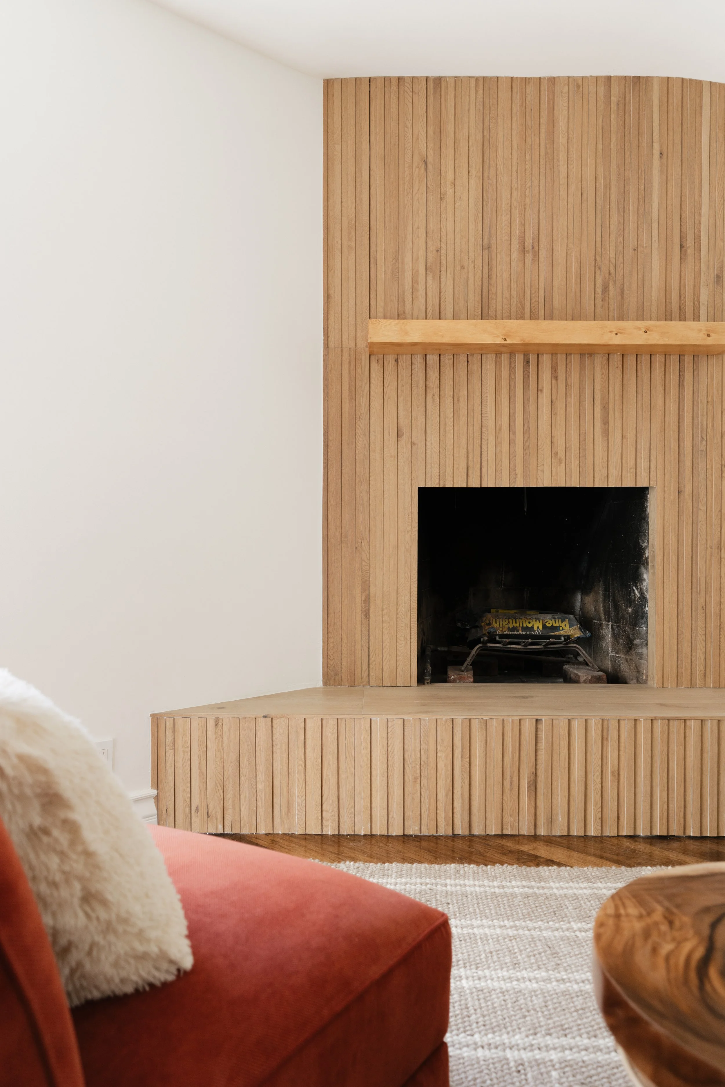 A living room with a wooden fireplace surround and mantel, with a red sofa and cream pillow, and a hardwood floor and area rug.