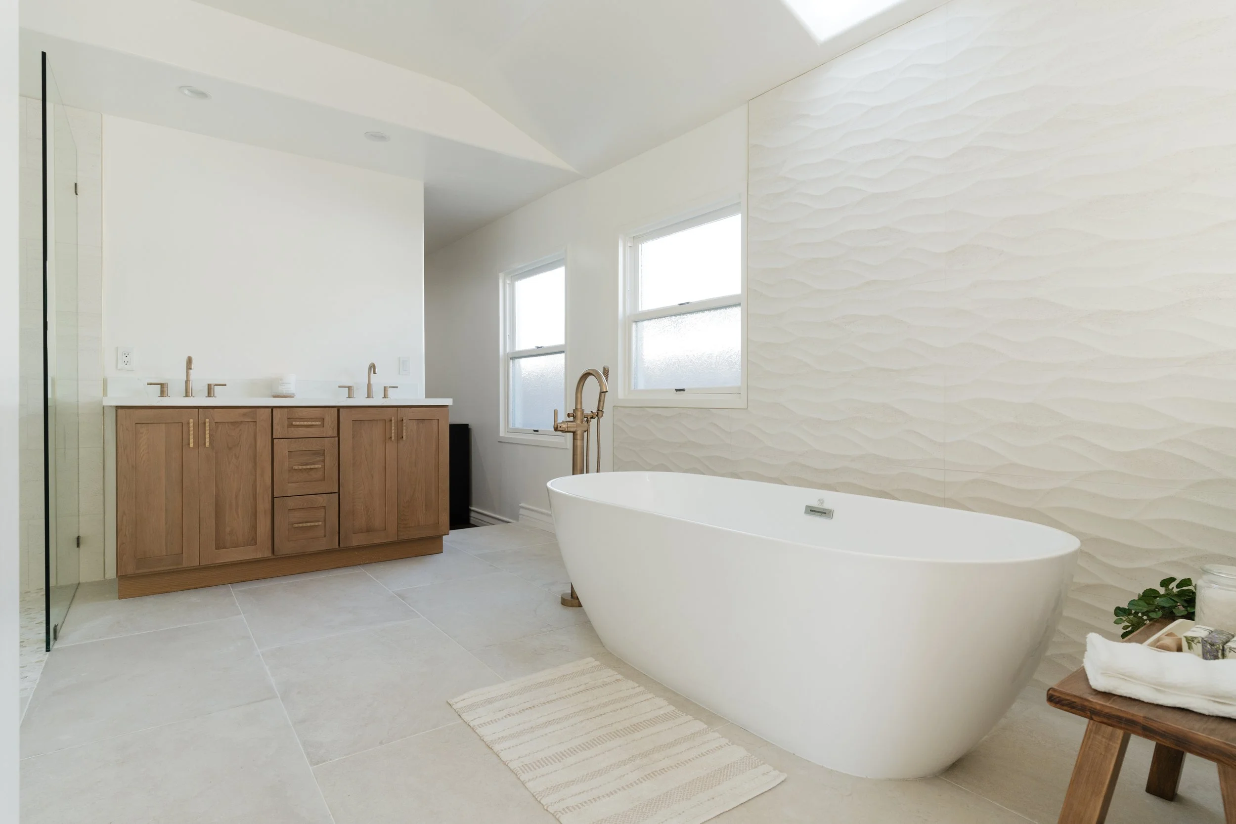 Modern bathroom with a freestanding bathtub with freestanding champagne bronze tub filler, wooden vanity with two sinks, textured white wall, and two windows.