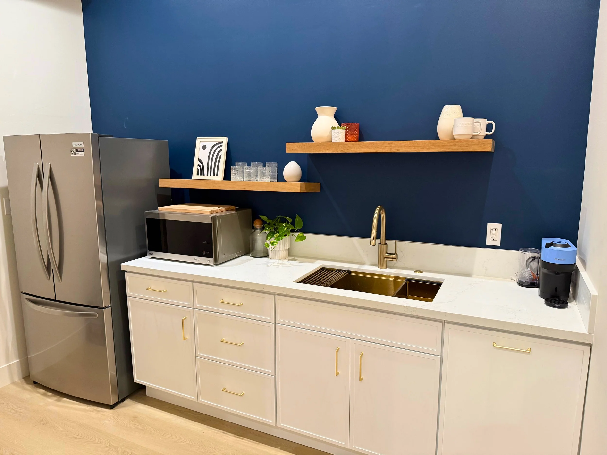 Kitchen with a stainless steel refrigerator, white cabinets with gold handles, a white countertop with a gold sink, a microwave, potted plant, and open wooden shelves with decorative vases and cups against a dark blue accent wall.