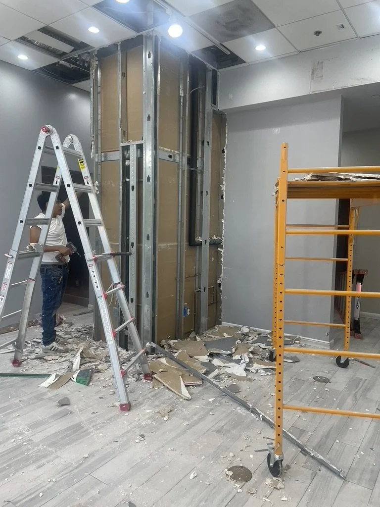Construction worker doing a full remodel of a commercial space, standing next to a ladder inside a room with damaged drywall and a metal framework on the wall.