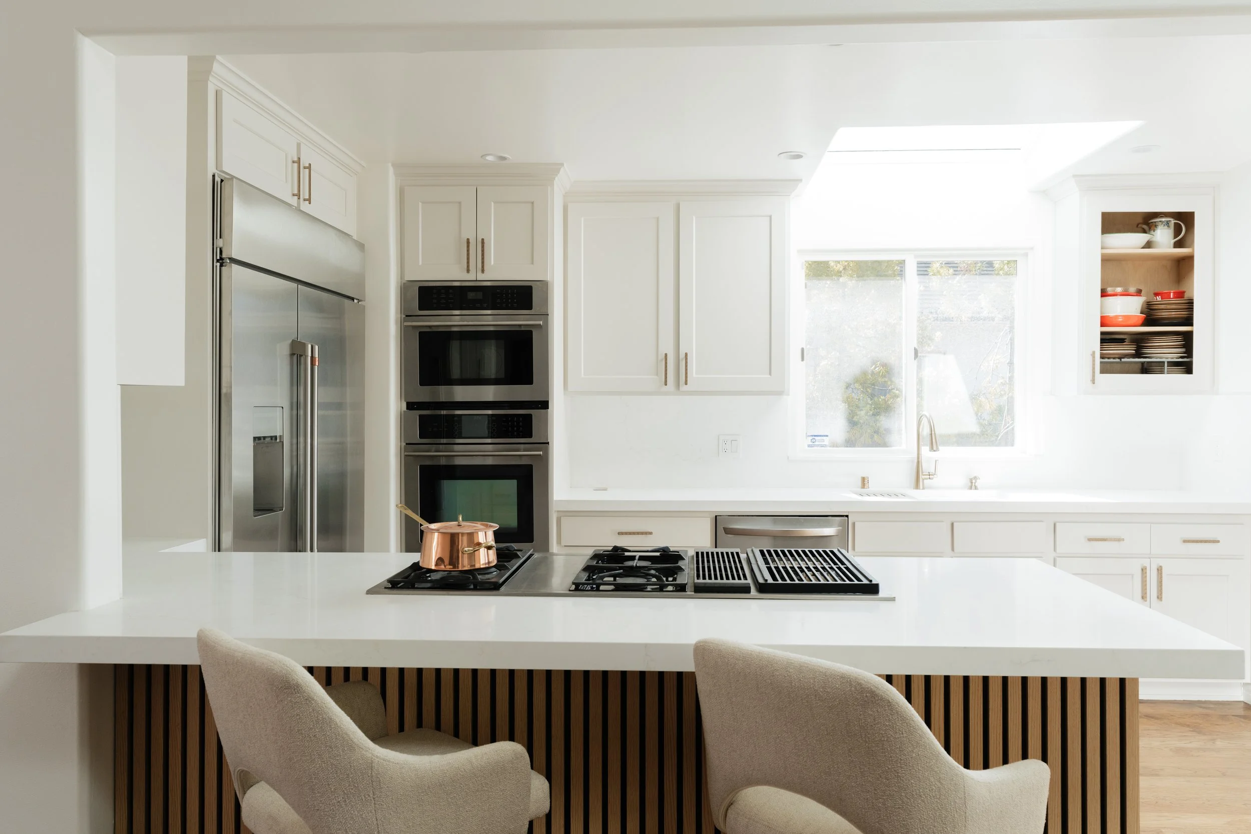 Modern white kitchen with island, copper pot on stove, built-in oven, refrigerator, open shelf with dishes, window above sink.