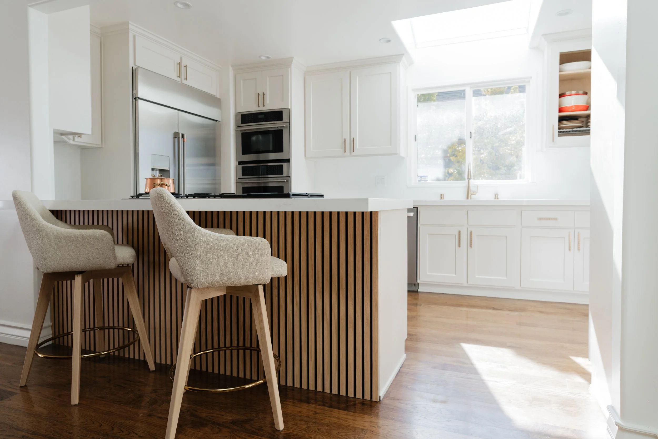 Modern white kitchen with a breakfast bar and beige barstools, stainless steel appliances, and a large window bringing in natural light.
