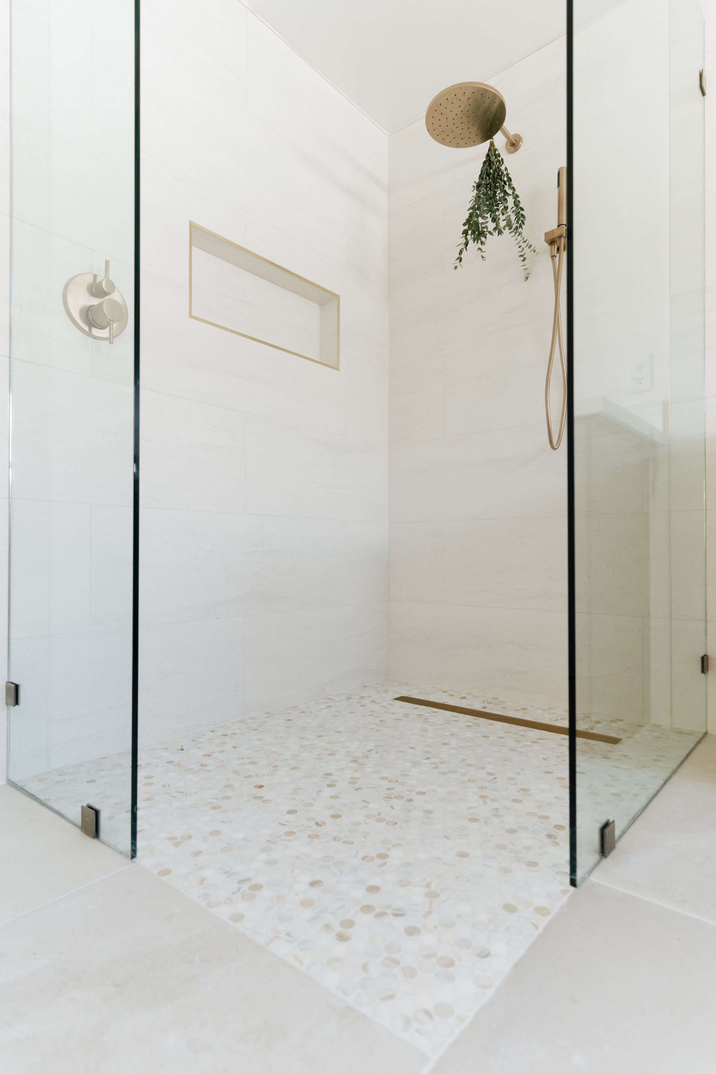Minimalist walk-in shower with a round rainfall shower head, a built-in wall niche, and a hanging plant, enclosed by clear glass panels.
