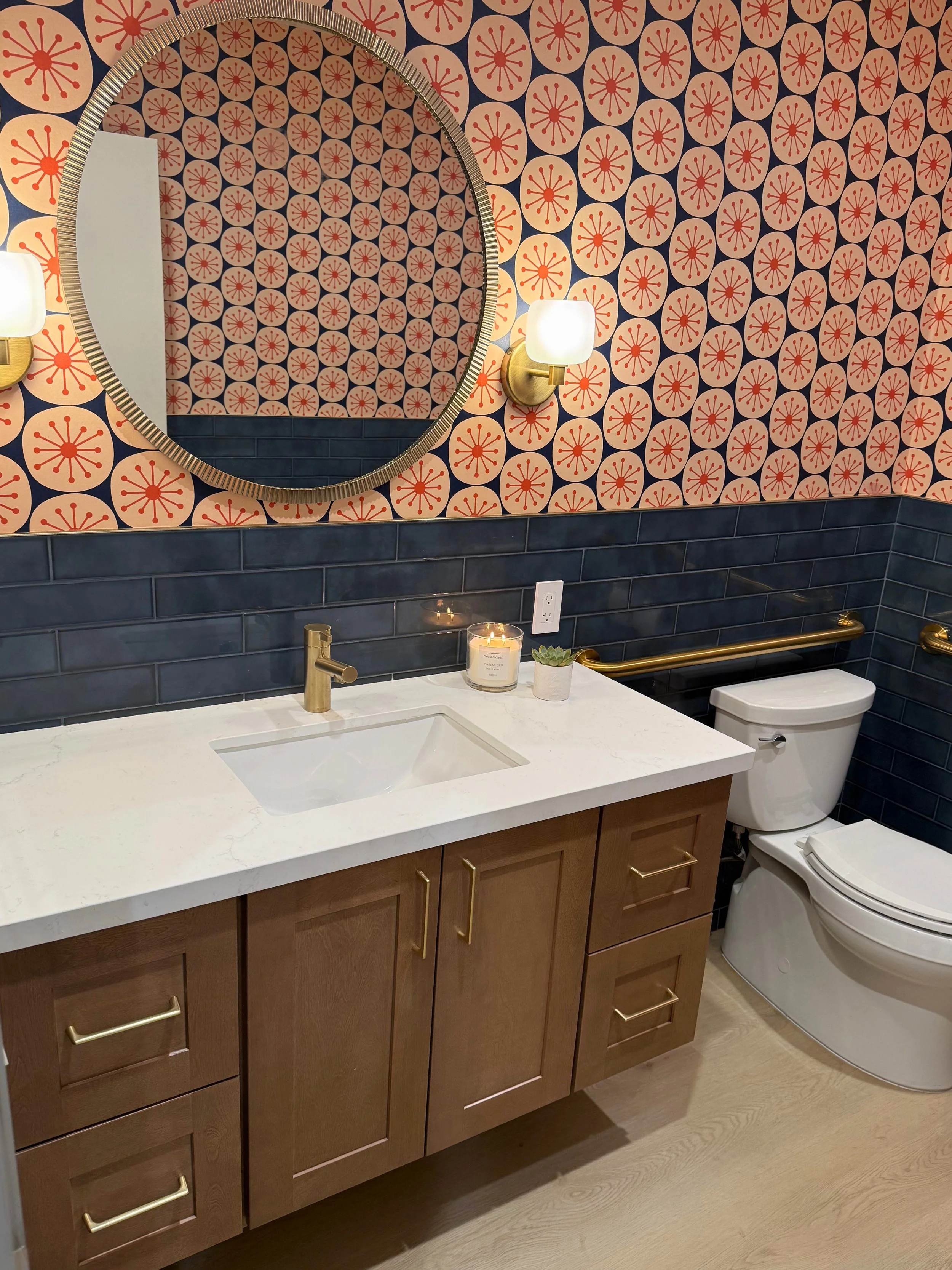 A modern bathroom with a wooden vanity, white countertop, gold fixtures, dark blue tile walls, a round mirror, wall sconces, a candle, and a toilet.